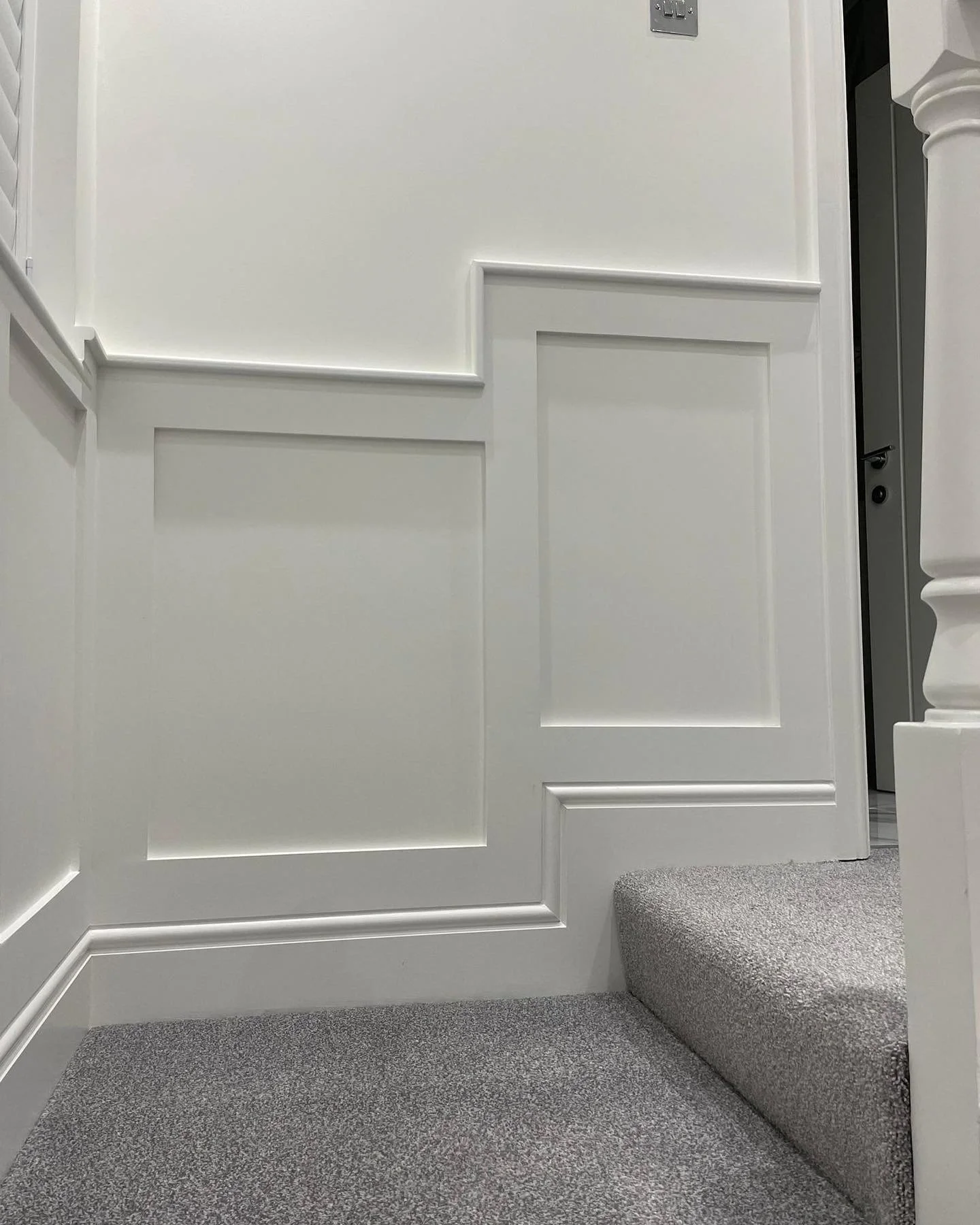 Close-up view of a white interior door with paneled design, a small set of stairs with gray carpeted steps to the right, and a section of a gray carpeted floor in a well-lit room.