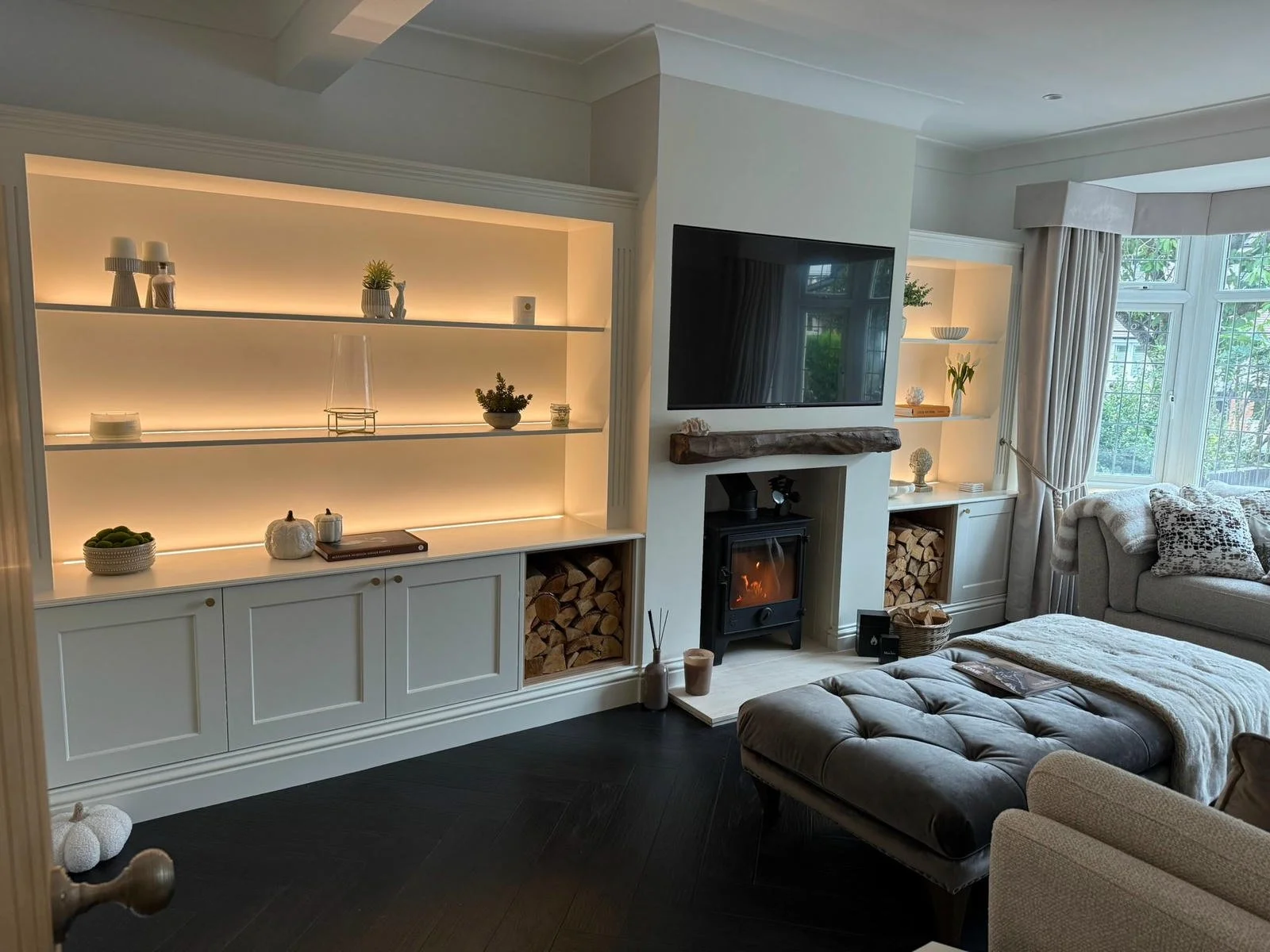Living room with white built-in shelving units, a flat-screen TV, and a fireplace with stacked firewood underneath. A gray sofa with patterned pillows, a tufted ottoman, and large windows with cream curtains are present. Decorative items like vases, 