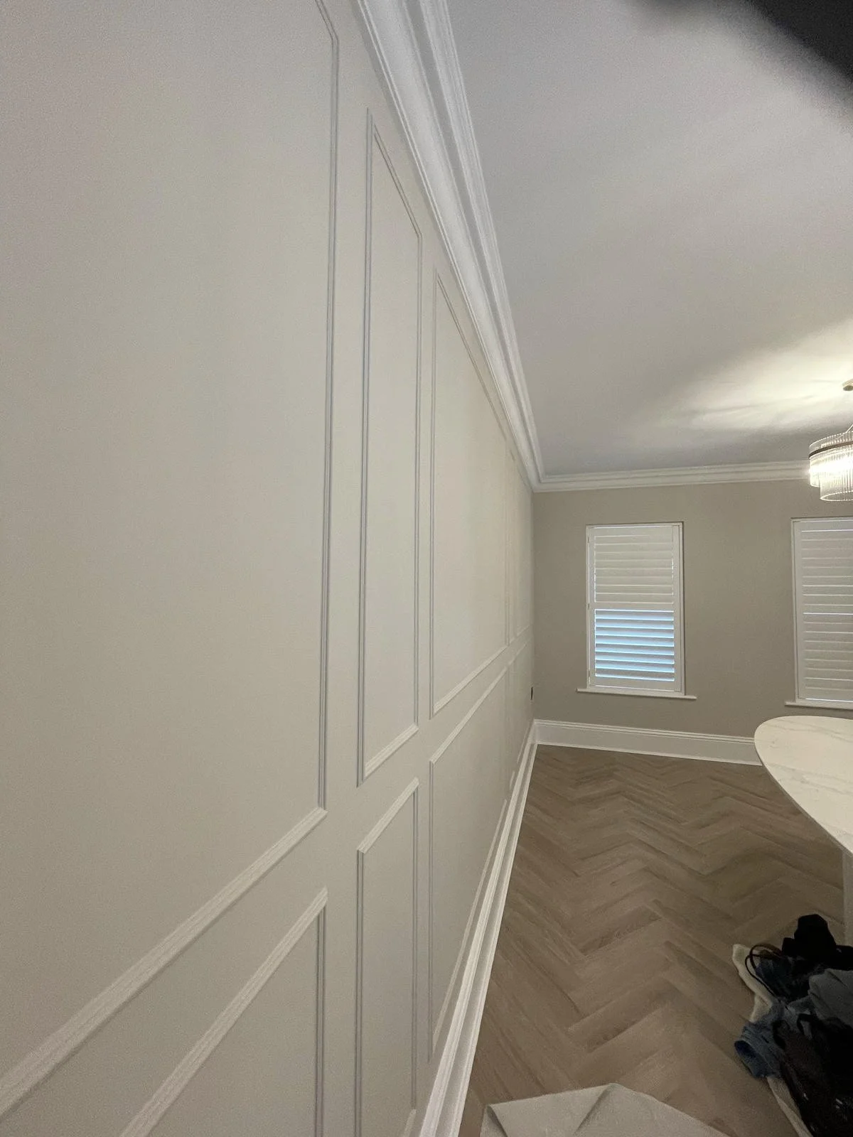 Interior of a room with beige walls featuring decorative molding, three windows with blinds, a chandelier, and hardwood herringbone flooring.