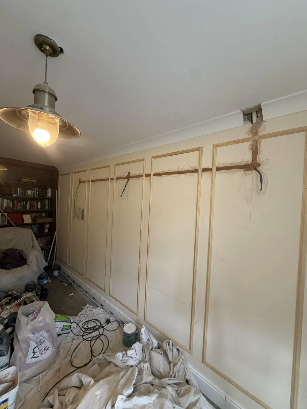 A room under renovation with exposed wall framing, construction tools, paint cans, and a cluttered floor. There is a ceiling light fixture and a bookshelf filled with books.