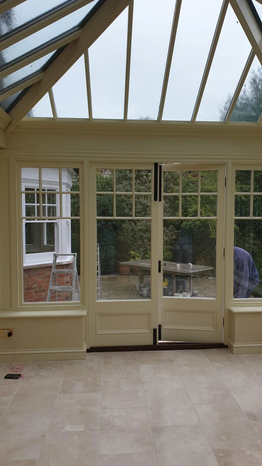 View from inside a conservatory with large glass doors and upper window panes, overlooking a patio area with a table, potted plants, and two people working outside.