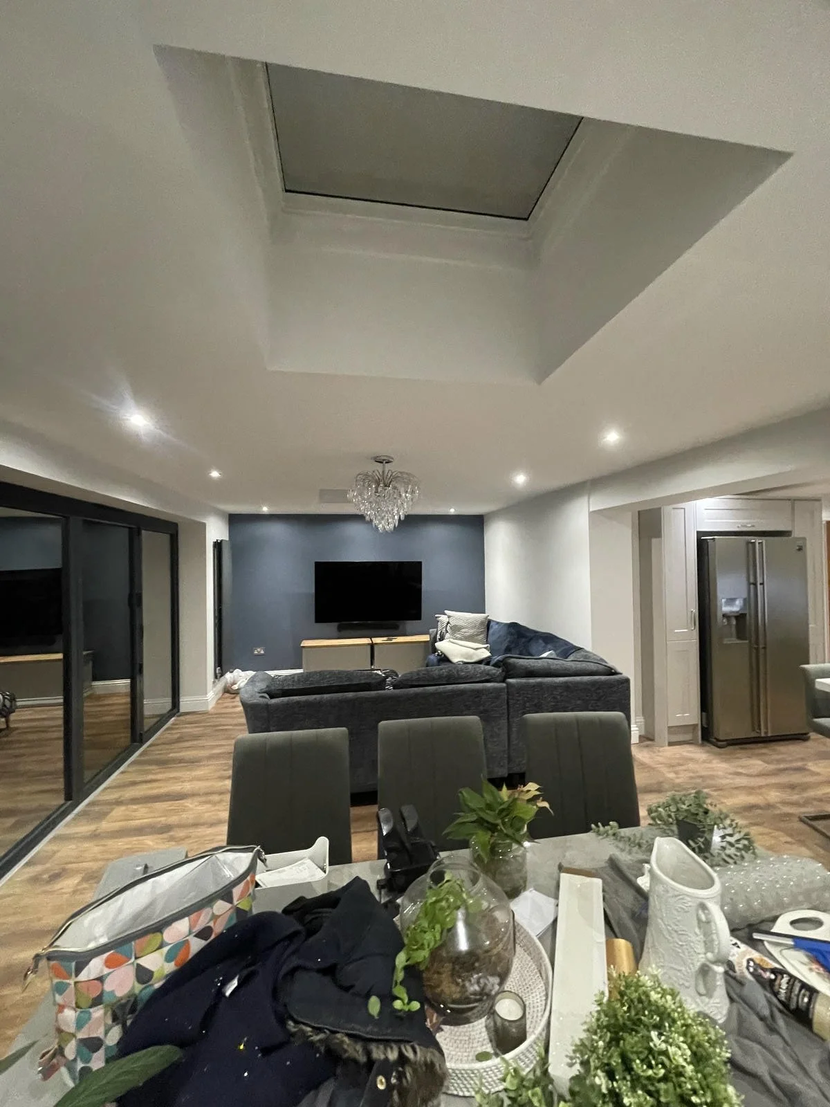 Living room with hardwood floors, dark blue accent wall, television, gray sofa, chandelier, sitting area, kitchen with refrigerator, and cluttered dining table.