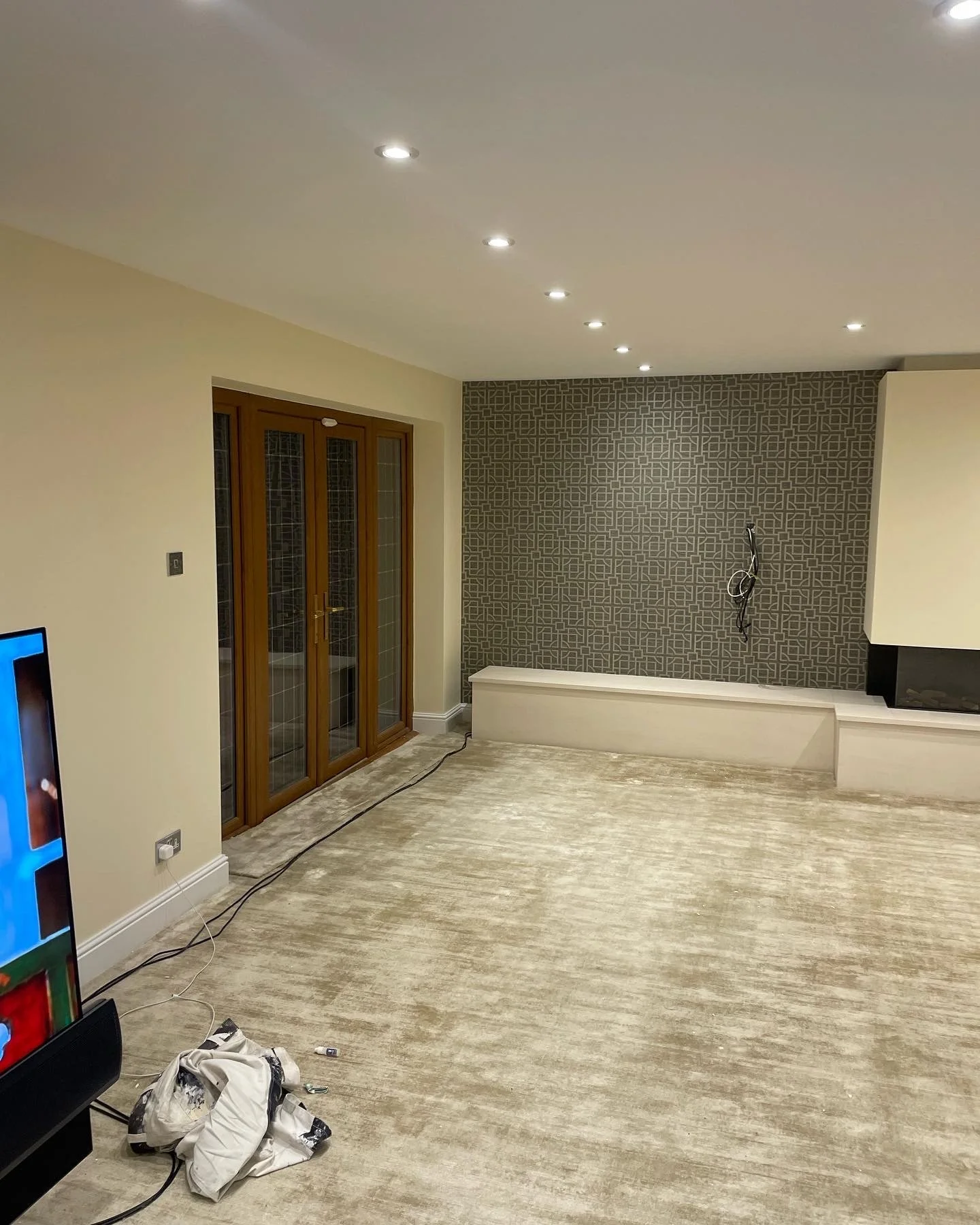 Living room with a carpeted floor, partially installed, with a wall featuring geometric patterned wallpaper, a fireplace, and a set of wooden glass-paneled double doors. There are ceiling spotlights and some electrical wires hanging on the wall and floor.
