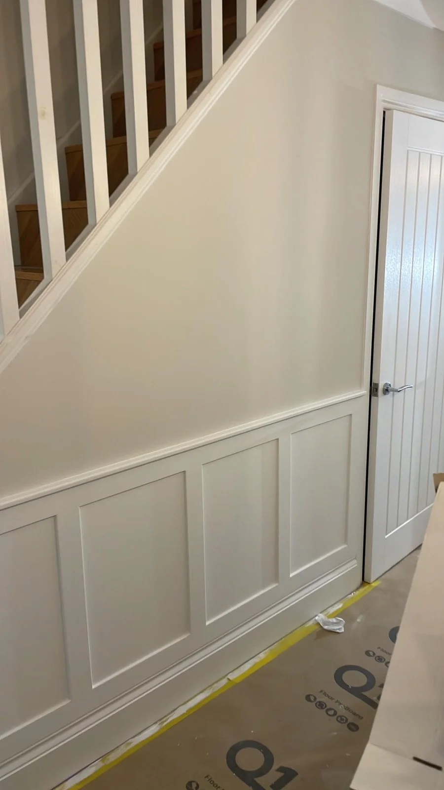 Interior view of a stairwell with white wainscoting and a sloped wall, white paneled door, and wooden stairs with white railing visible at the top.