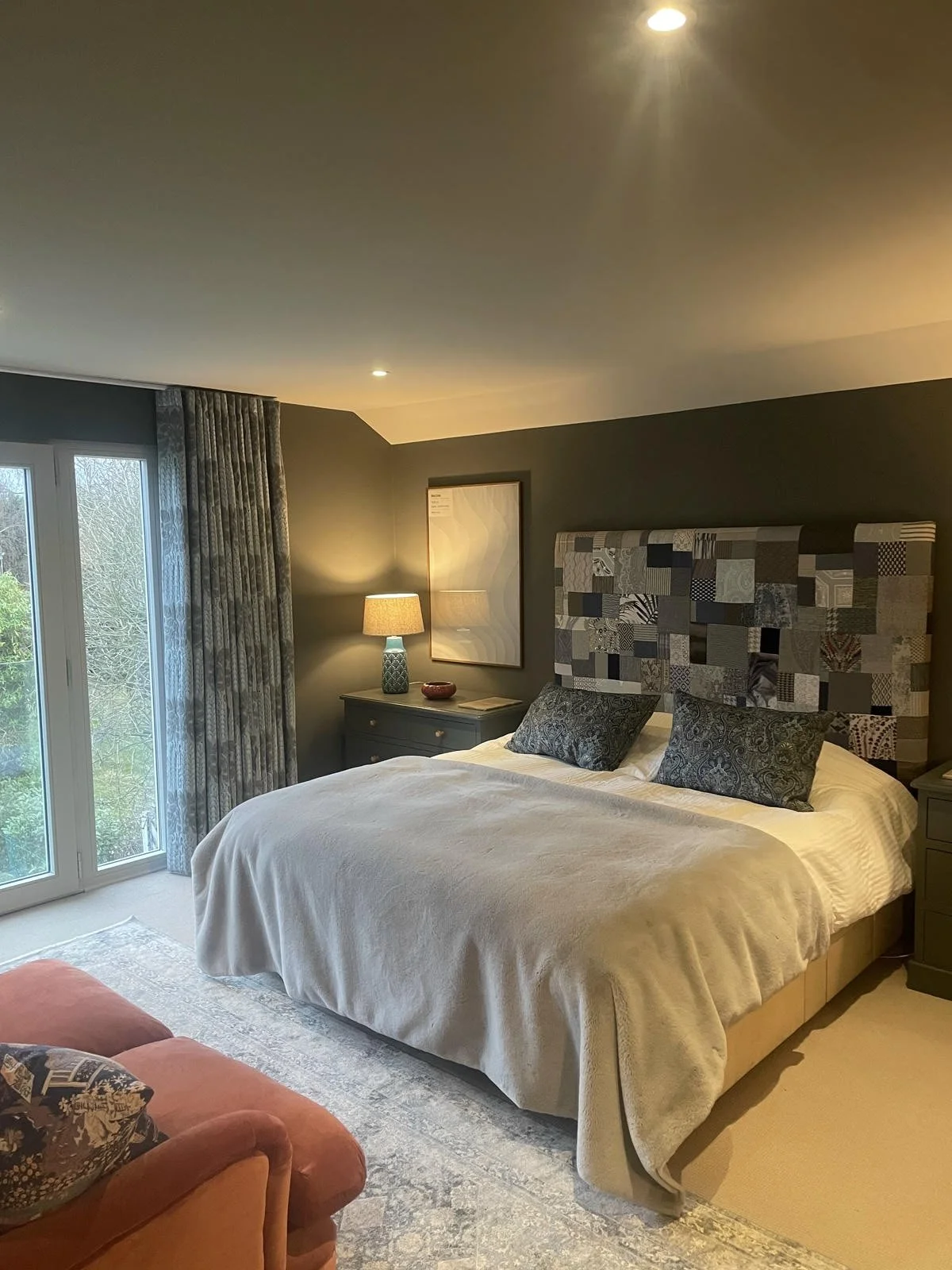 A bedroom with a large bed featuring a patchwork fabric headboard, two patterned pillows, and a beige bedspread. To the left, there are glass sliding doors with patterned curtains, revealing green trees outside. Next to the bed, a nightstand with a table lamp, a framed art piece, and a small decorative bowl. An orange armchair with a patterned pillow is partially visible in the foreground.
