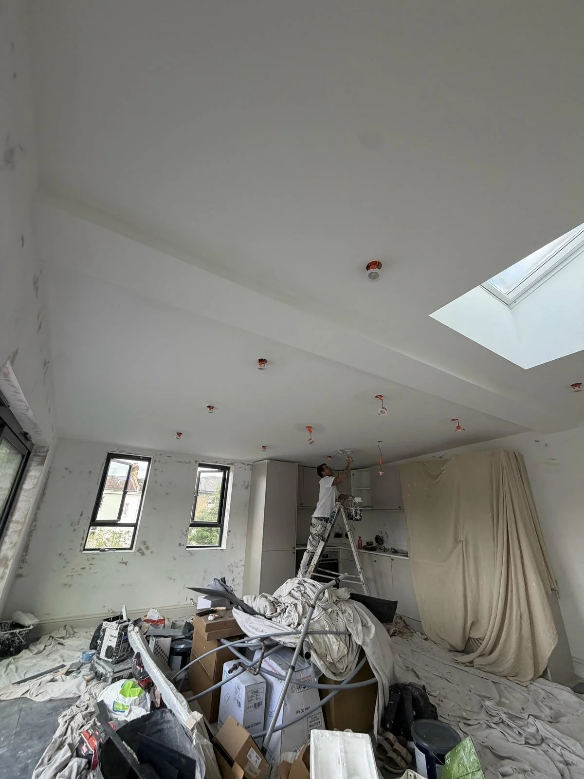 A construction worker standing on a ladder working on the ceiling of a room. The room is cluttered with tools, boxes, and construction materials, with multiple windows and a skylight.