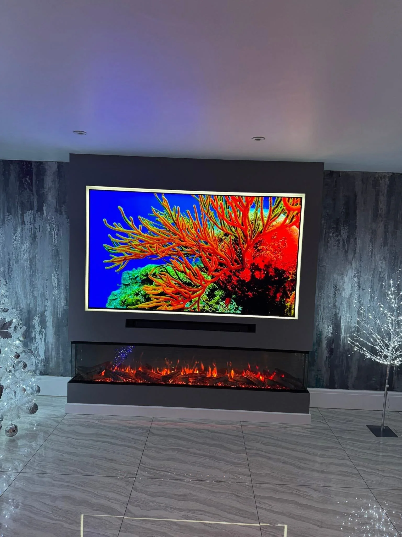 A wall-mounted flat-screen television displaying a colorful image of coral reef and fish, with a modern electric fireplace below it emitting orange and red flames, in a contemporary living room with gray textured walls and glossy marble flooring.