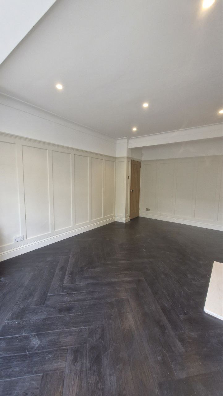 Empty room with dark wood herringbone flooring, white paneled walls, and ceiling with recessed lighting.