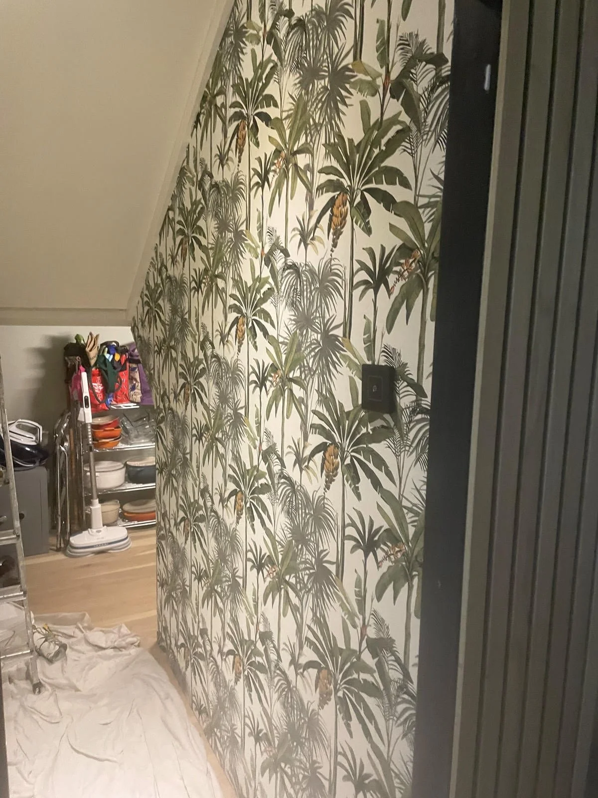 Interior of a room with tropical-themed wallpaper featuring green leaves and yellow-orange flowers. There is a black door or panel on the right side, and a small shelf with cleaning supplies and kitchen items on the left. The floor has a white sheet 
