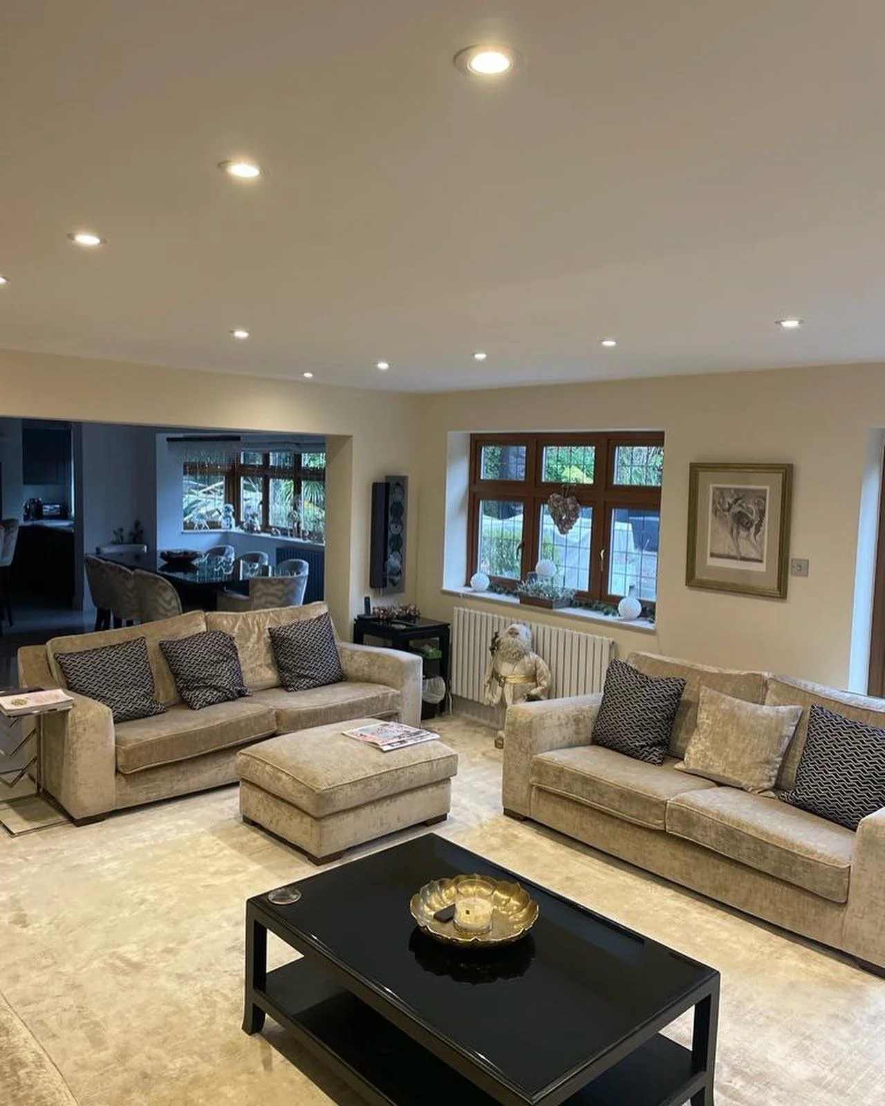 Living room with beige sofas, pillows, and a black coffee table, decorated with framed artwork, a window with a view of greenery, and a cozy ambiance.