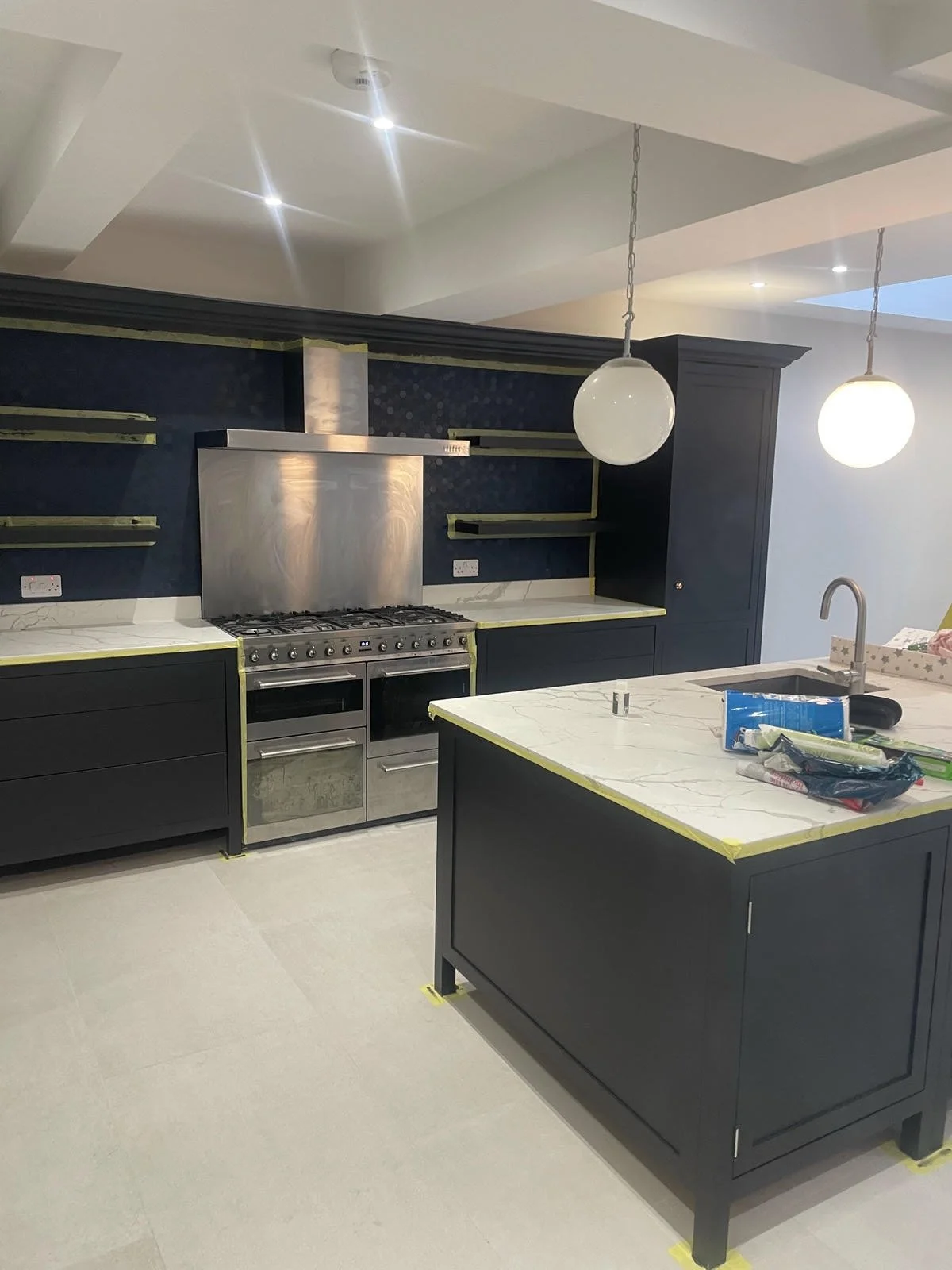 Modern kitchen with dark blue cabinets, stainless steel stove and range hood, marble countertops, and pendant lighting. Some items are on the island counter, and the cabinets and shelves are still under installation.
