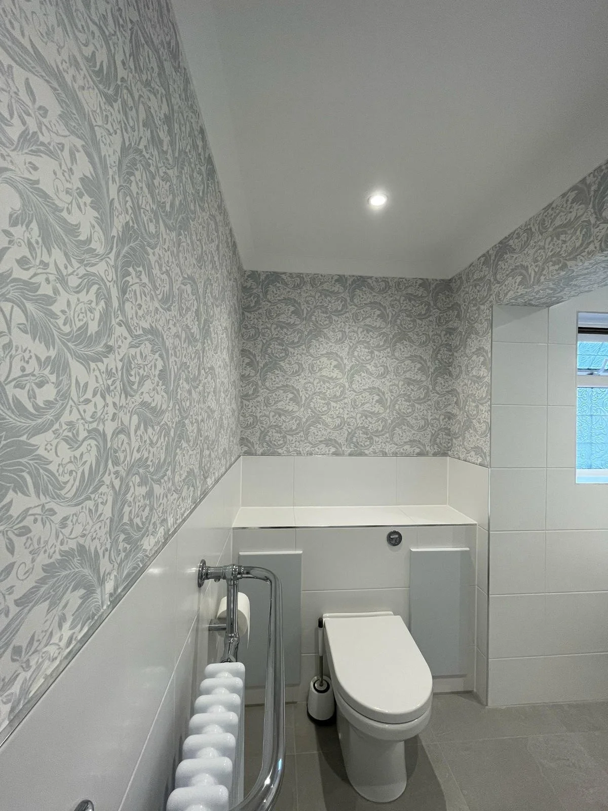 Bathroom with floral wallpaper on the wall behind a toilet, white tiling, and a small window with blue patterned glass.