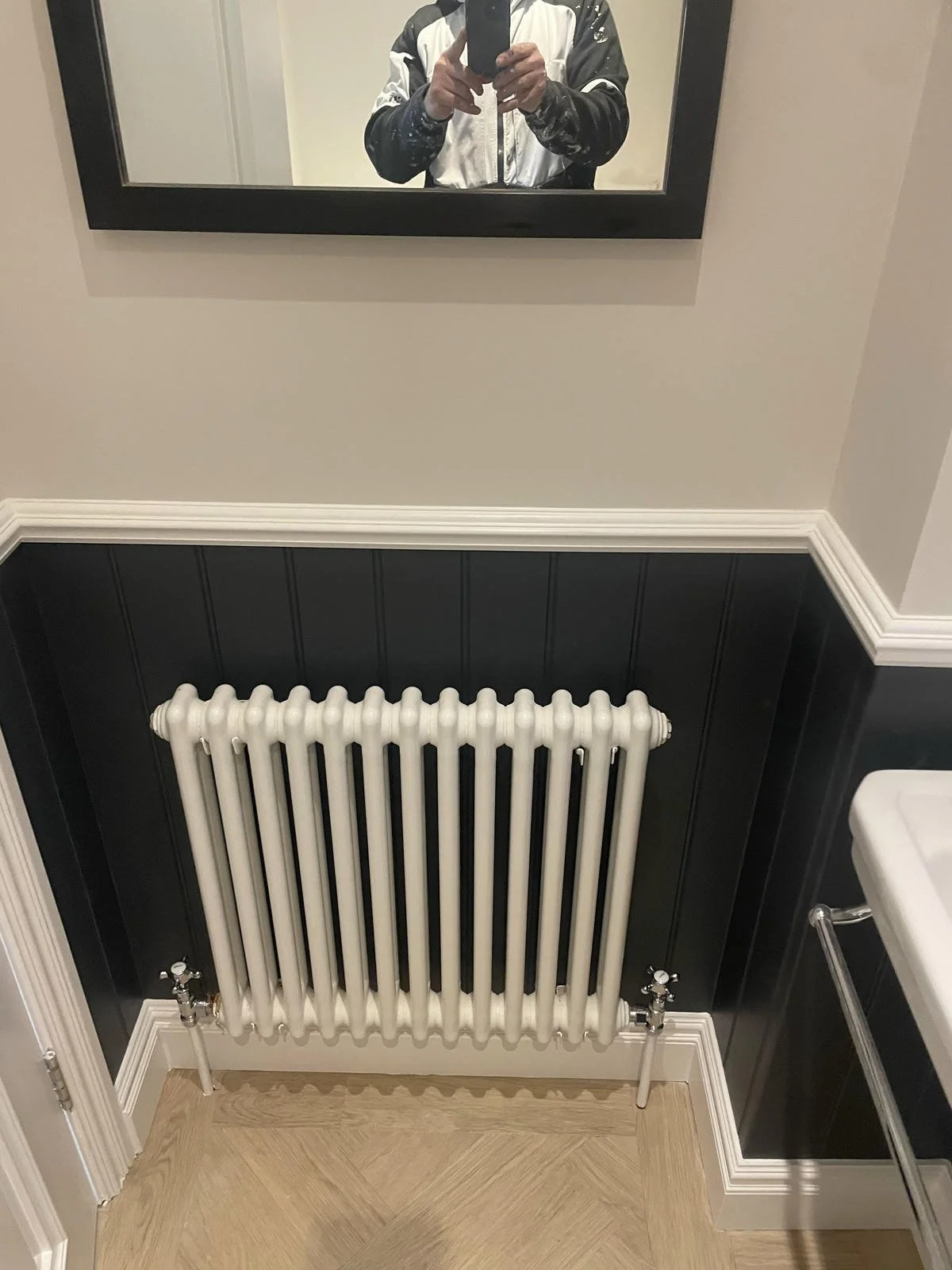 A white vintage radiator centered against a black wainscoting wall with a white wall above. A mirror is visible at the top showing a person taking the photo. To the right, a portion of a white porcelain sink is seen. The room has light wooden floorin