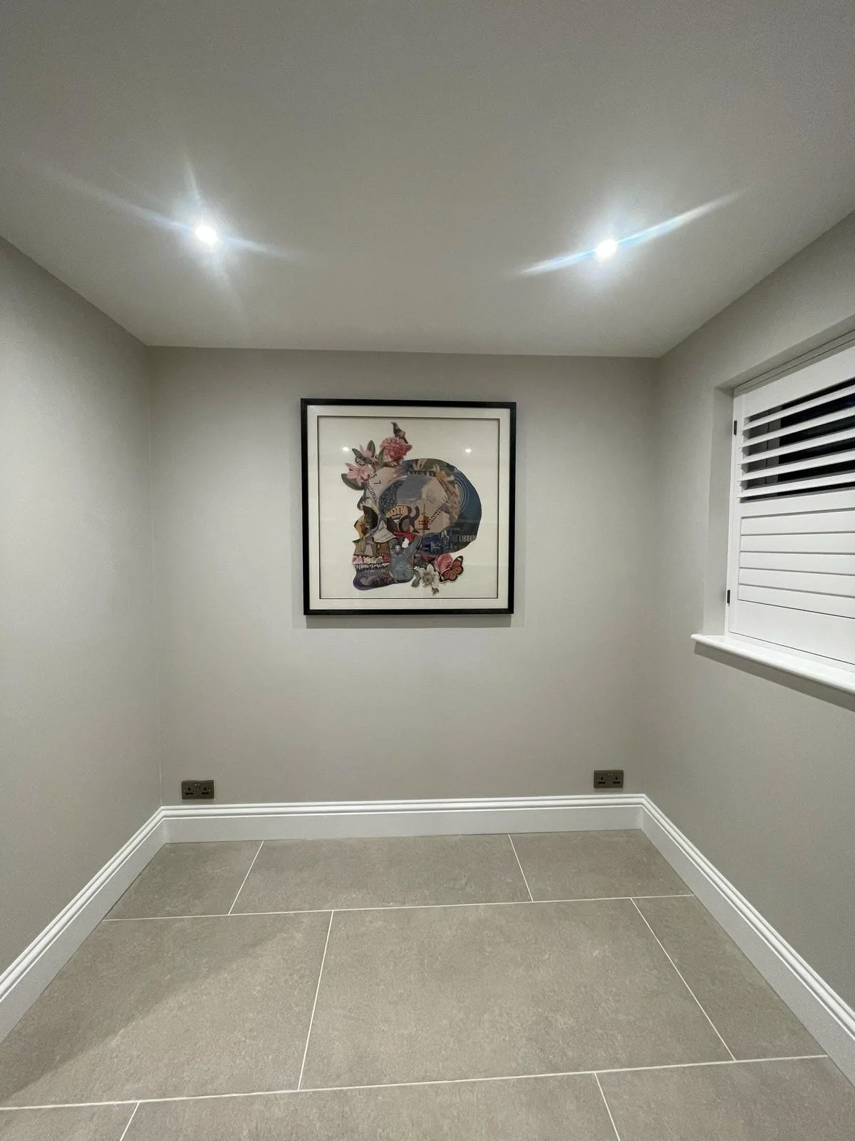 Empty room with gray walls, a framed colorful artwork of a skull with flowers, a window with blinds, and tiled floor.