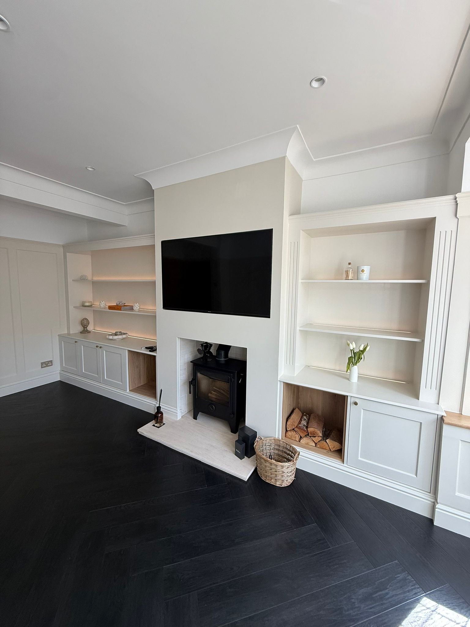 Living room with white built-in shelves, a black fireplace, a mounted flat-screen TV, and a basket of firewood, with dark hardwood floors.
