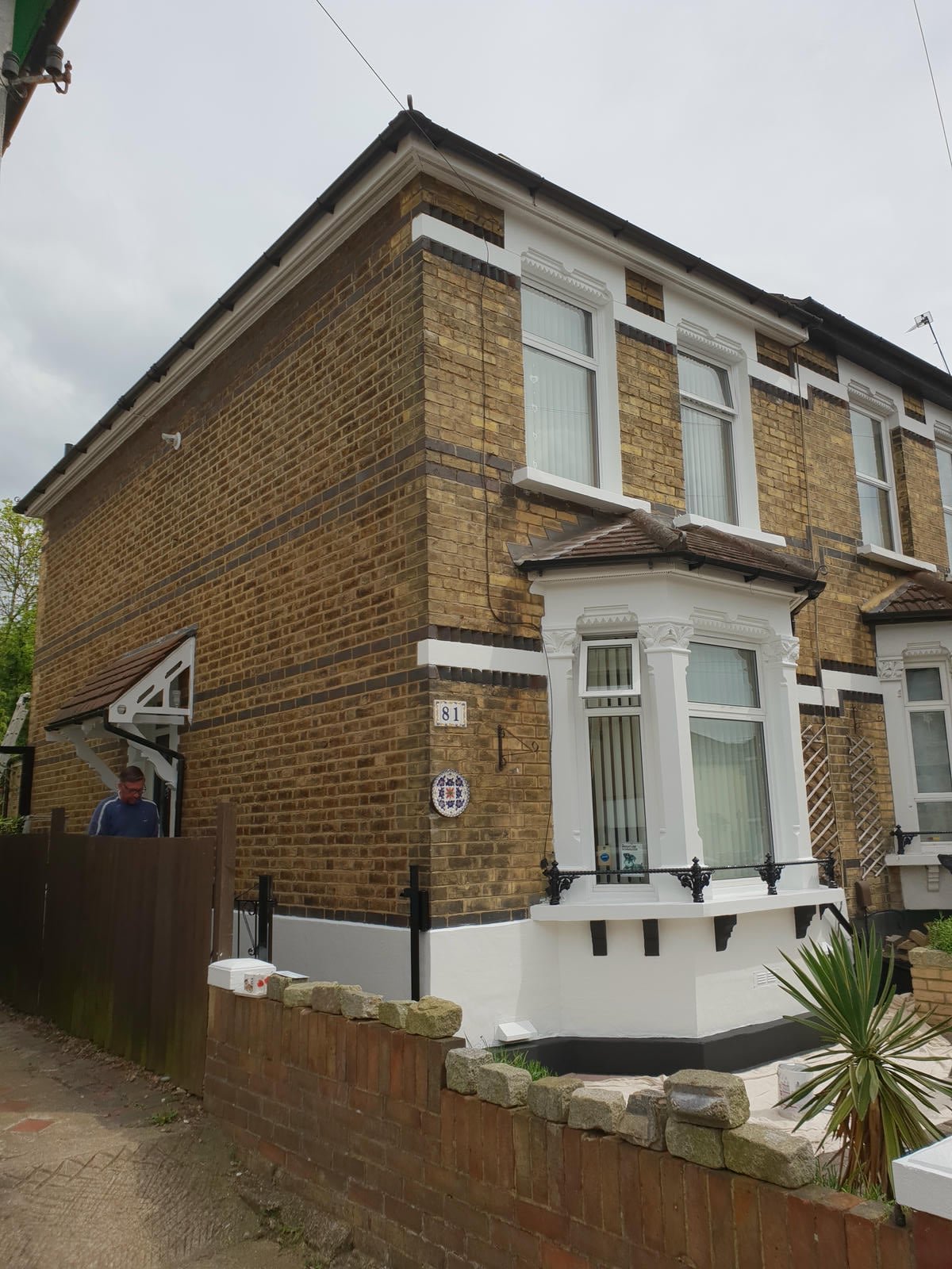 A traditional brick house with white decorative trim, bay windows, and a small front garden with a brick wall and a person standing near the entrance.