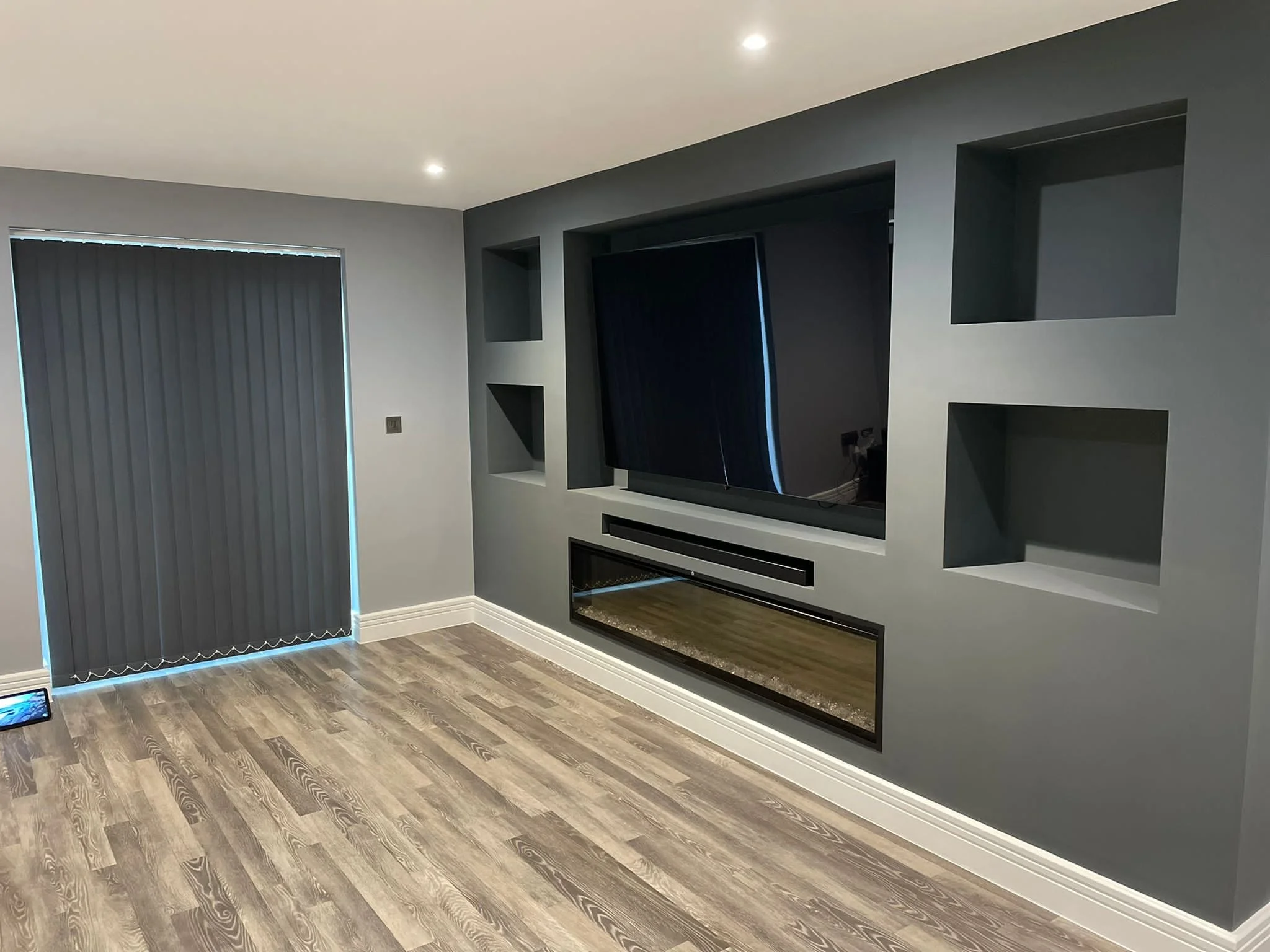 Living room with a flat-screen TV mounted on the wall, built-in shelves, gray walls, a window with closed gray vertical blinds, wooden flooring, and recessed ceiling lights.