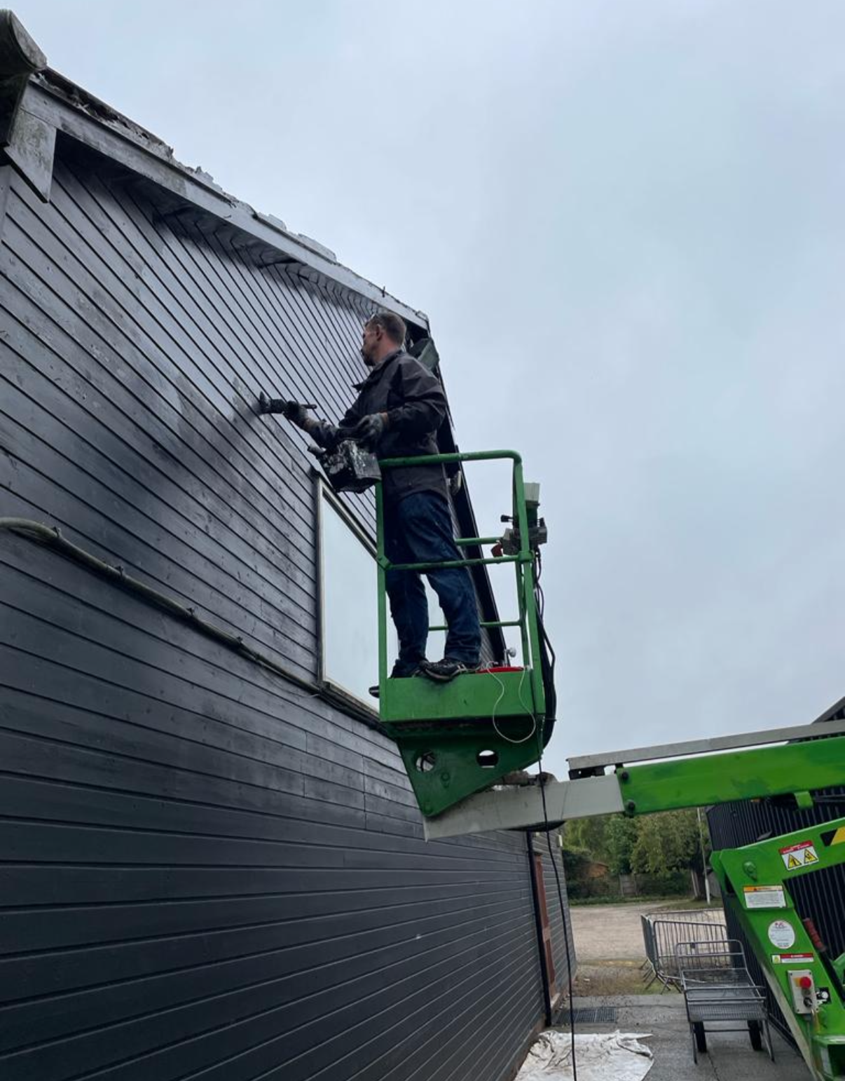 A man in a black jacket and jeans is standing on a green lift platform, power washing the black exterior wall of a building with horizontal siding.