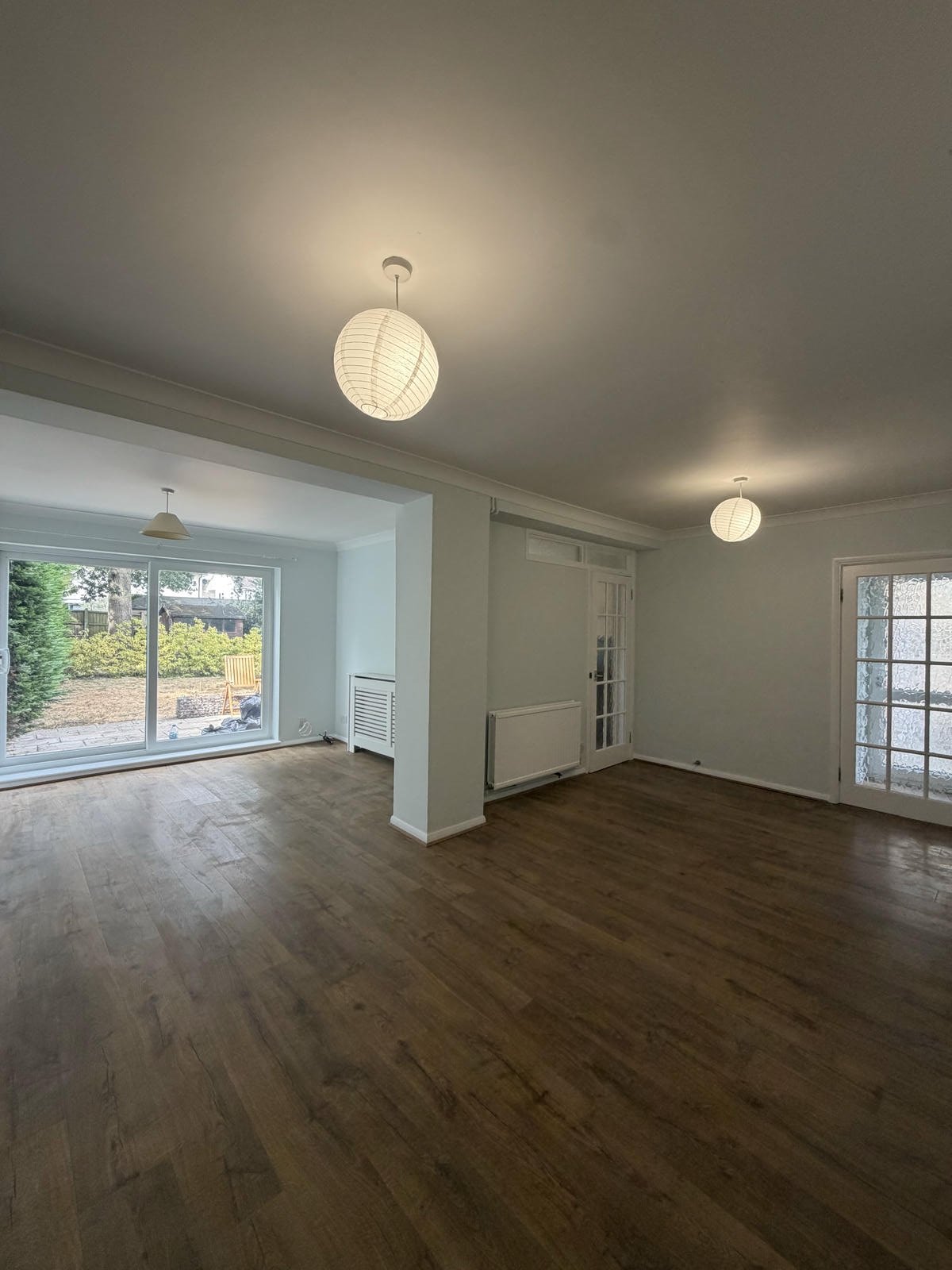 Empty living room with wooden floors, large windows, sliding glass door leading outside, and ceiling lights.