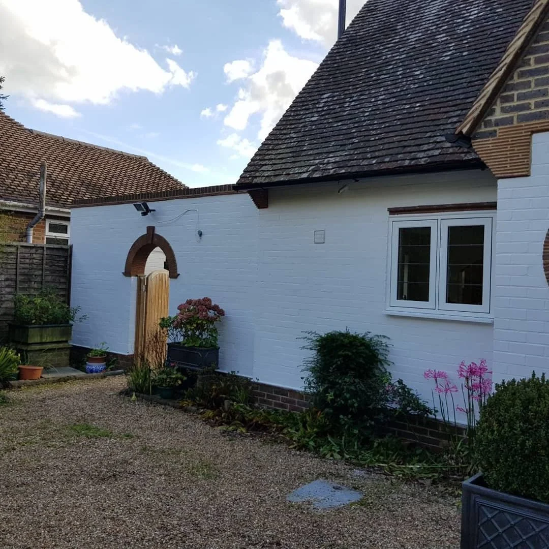A white brick house with a small window, a gravel patio, and a flower garden with pink and green plants. The house has a steep roof and an arched wooden gate leading to a side yard.