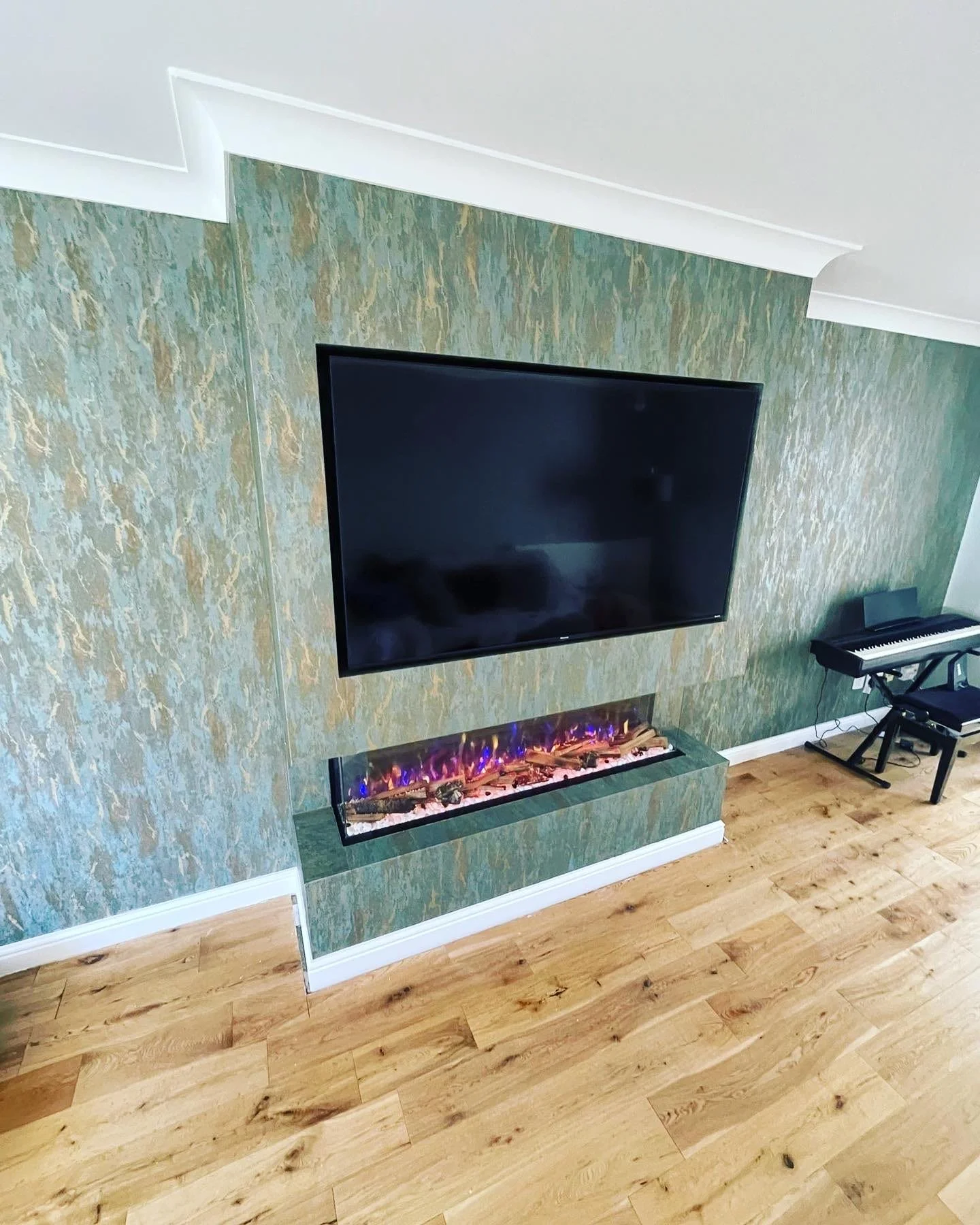 Living room with a flat-screen TV mounted on a green marble accent wall, electric fireplace below the TV, keyboard on a stand to the right, and hardwood floors.