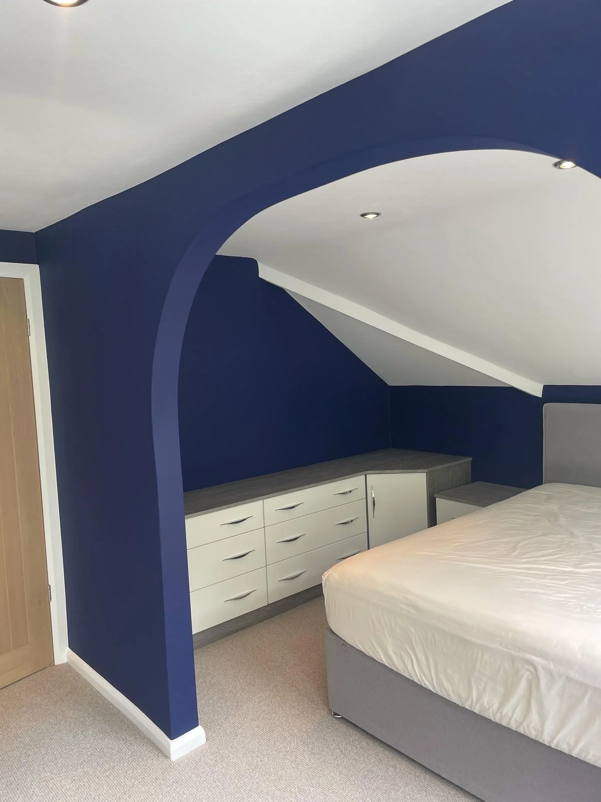 A bedroom with navy blue walls and ceiling, a white dresser with multiple drawers, a bed with a gray upholstered headboard, and recessed ceiling lights.