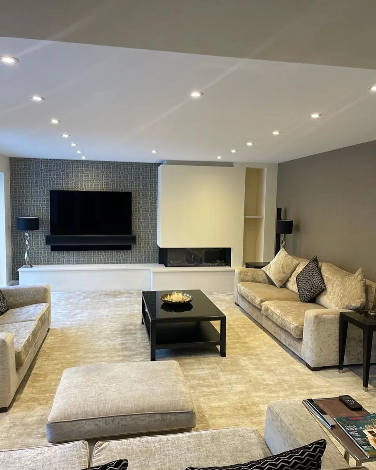 A modern living room with beige sofas, a black coffee table with a decorative bowl, a flat-screen TV on a grey patterned wall, and recessed ceiling lights.