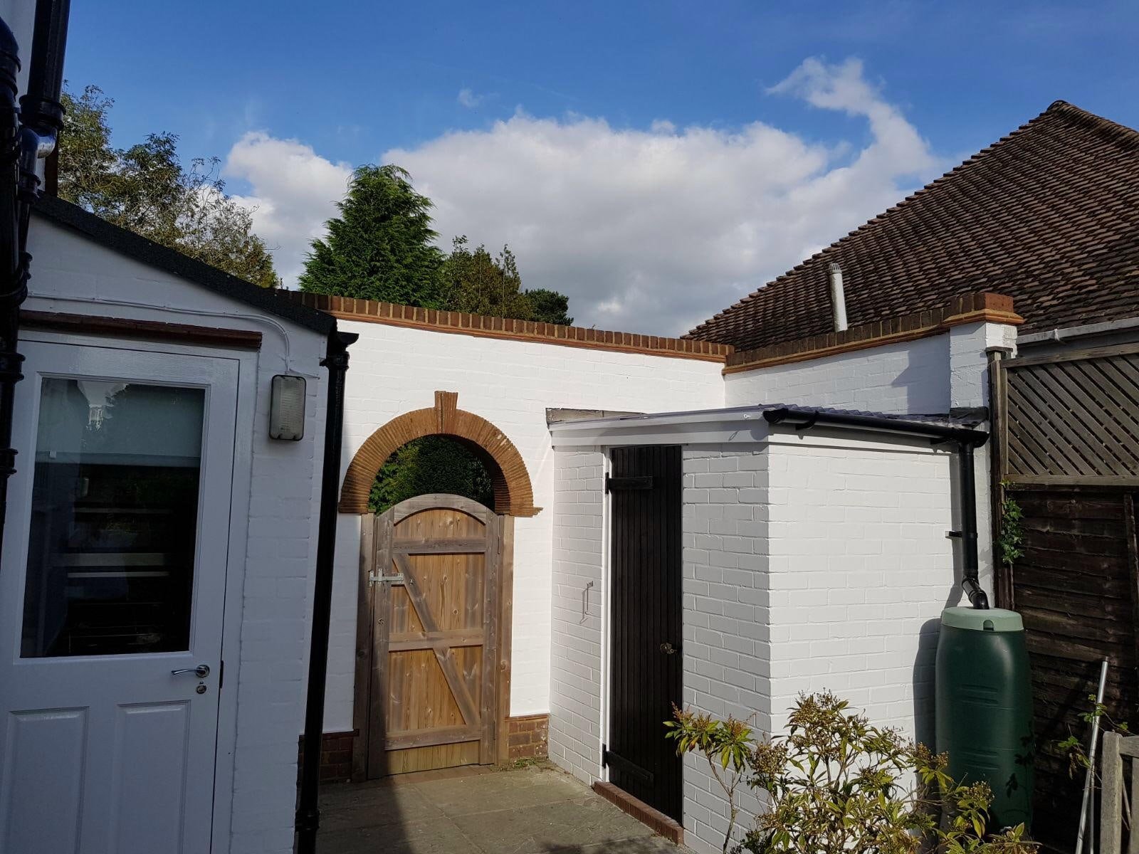 A backyard with a white brick wall, a wooden gate, a black metal door, a rainwater tank, and a wooden fence under a partly cloudy sky.