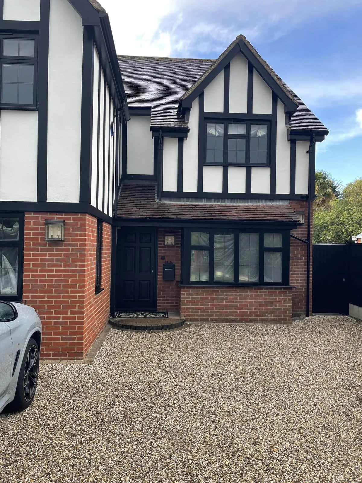 A two-story house with a brick ground floor and a white upper floor with black timber framing, featuring several windows and a black front door, with a gravel driveway and a parked car visible to the left.