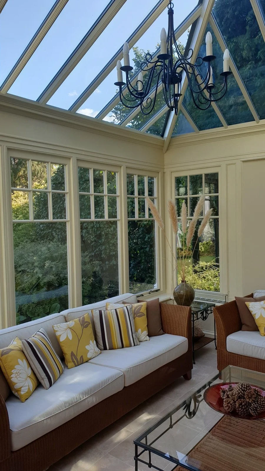 Sunroom with large windows, glass ceiling, and a black chandelier, furnished with a white and brown sofa, patterned pillows, a glass coffee table, and a vase with pampas grass.