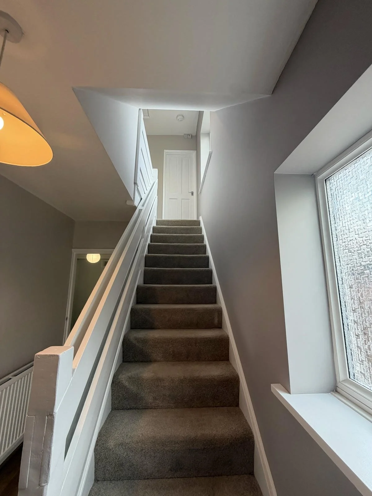 A carpeted staircase viewed from the bottom, leading to a closed white door at the top. The staircase is flanked by white railings and walls painted light gray. To the right, a large window with textured glass allows natural light into the area. On t