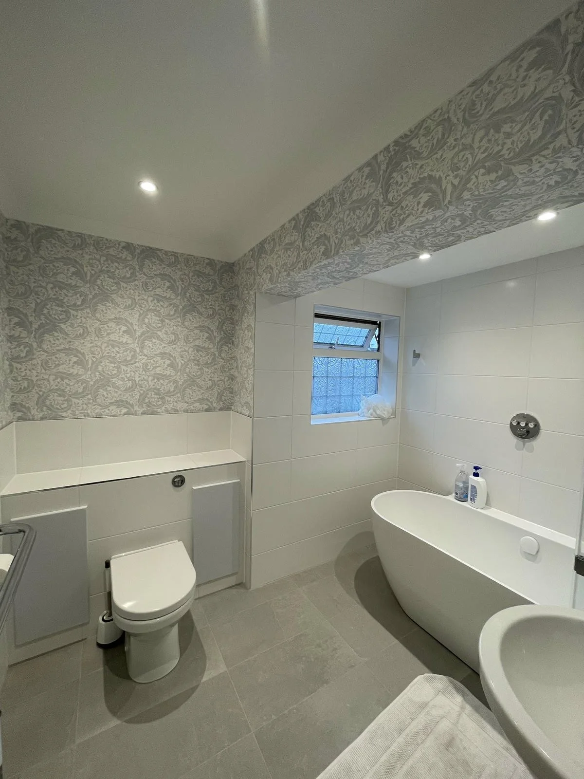 A modern bathroom with a freestanding bathtub, toilet, and a small window. The walls are tiled in white, and the ceiling features patterned wallpaper with recessed lighting.