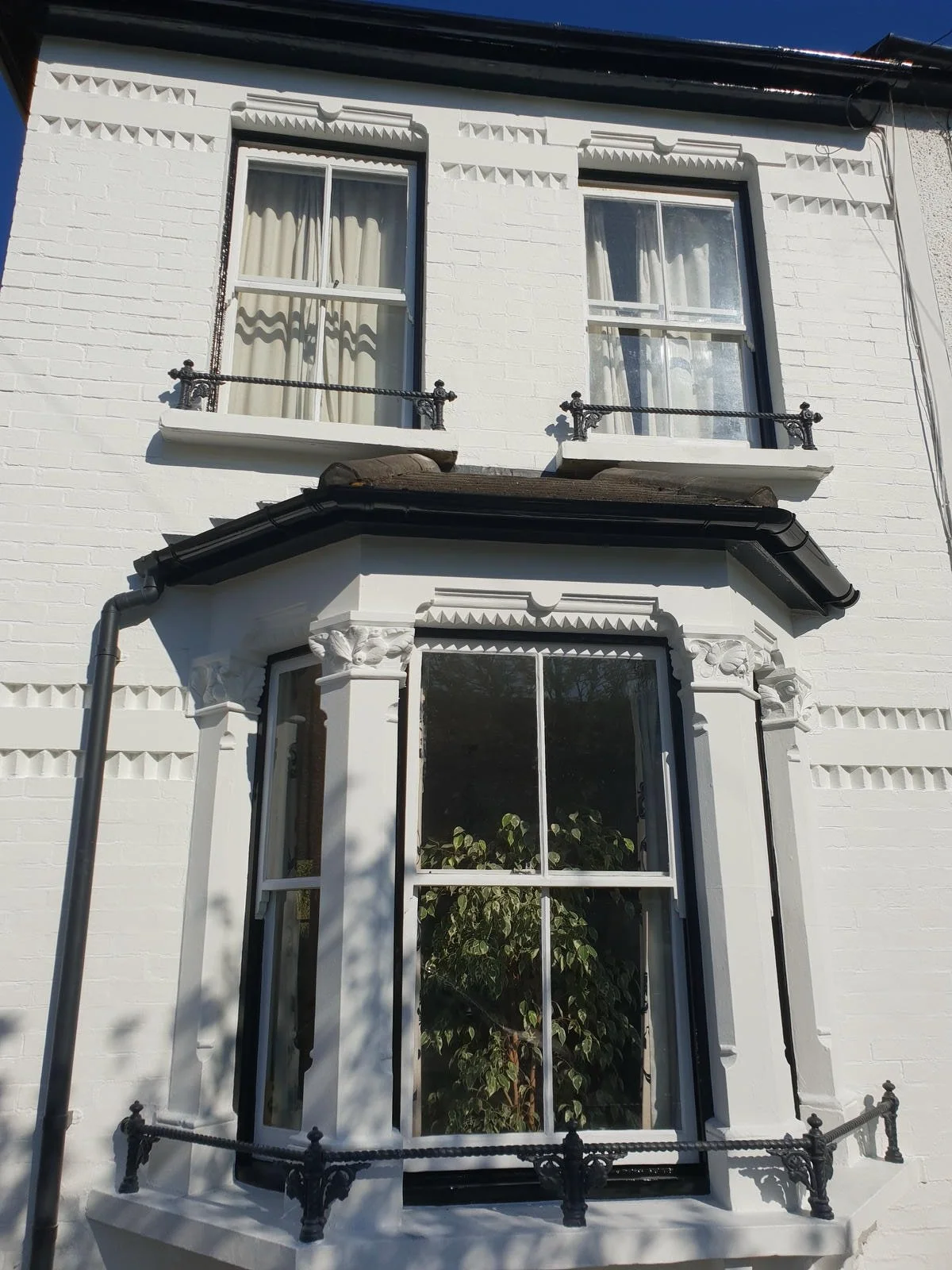 White brick house with large bay windows on the ground and second floors, decorative architectural details, black railings, and a black gutter downspout on the left side.