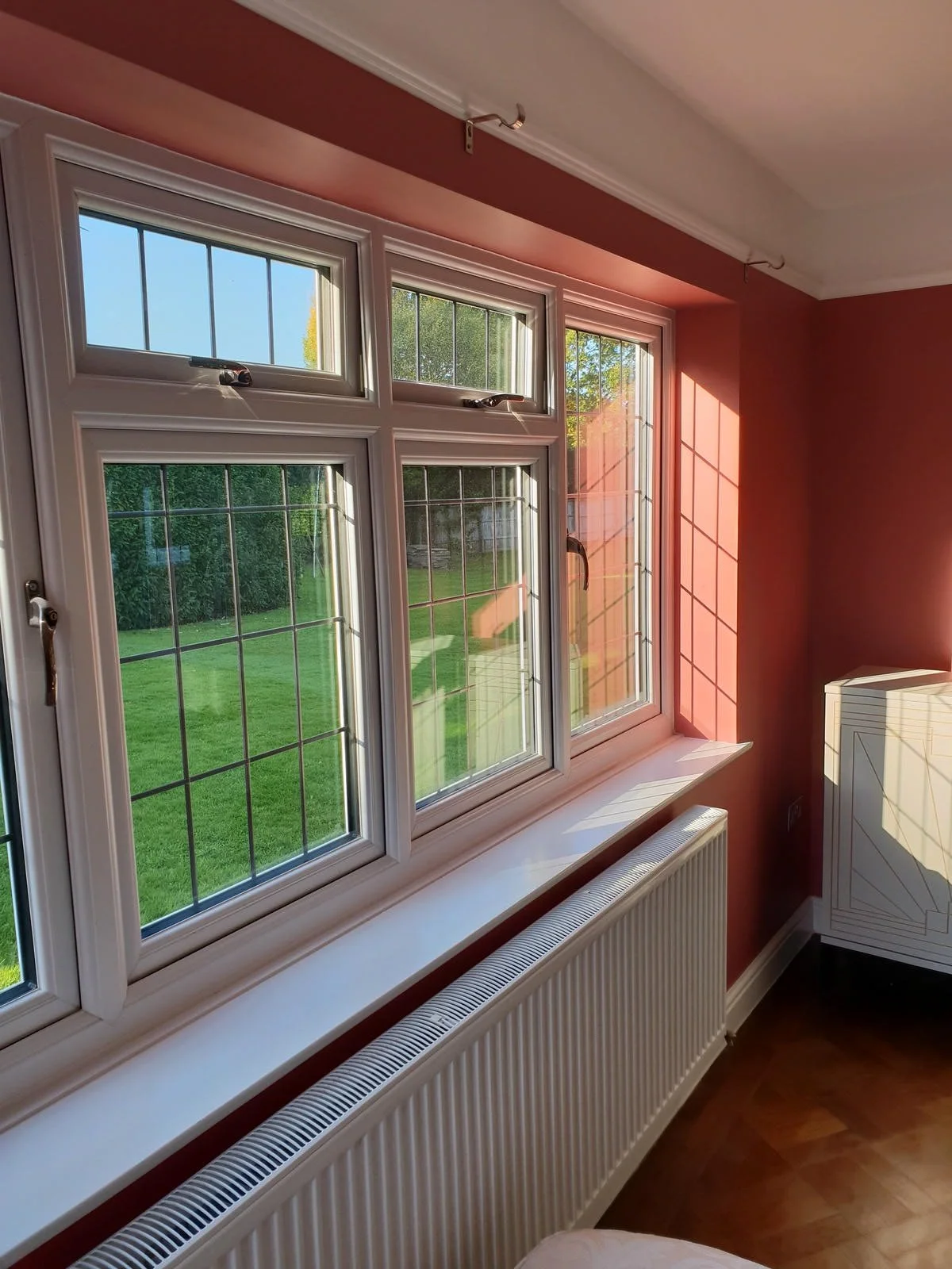 A room with a set of four white framed windows showing a green backyard with grass and trees, sunlight casting shadows inside, a white radiator below the windows, and a red wall with a white decorative corner piece.