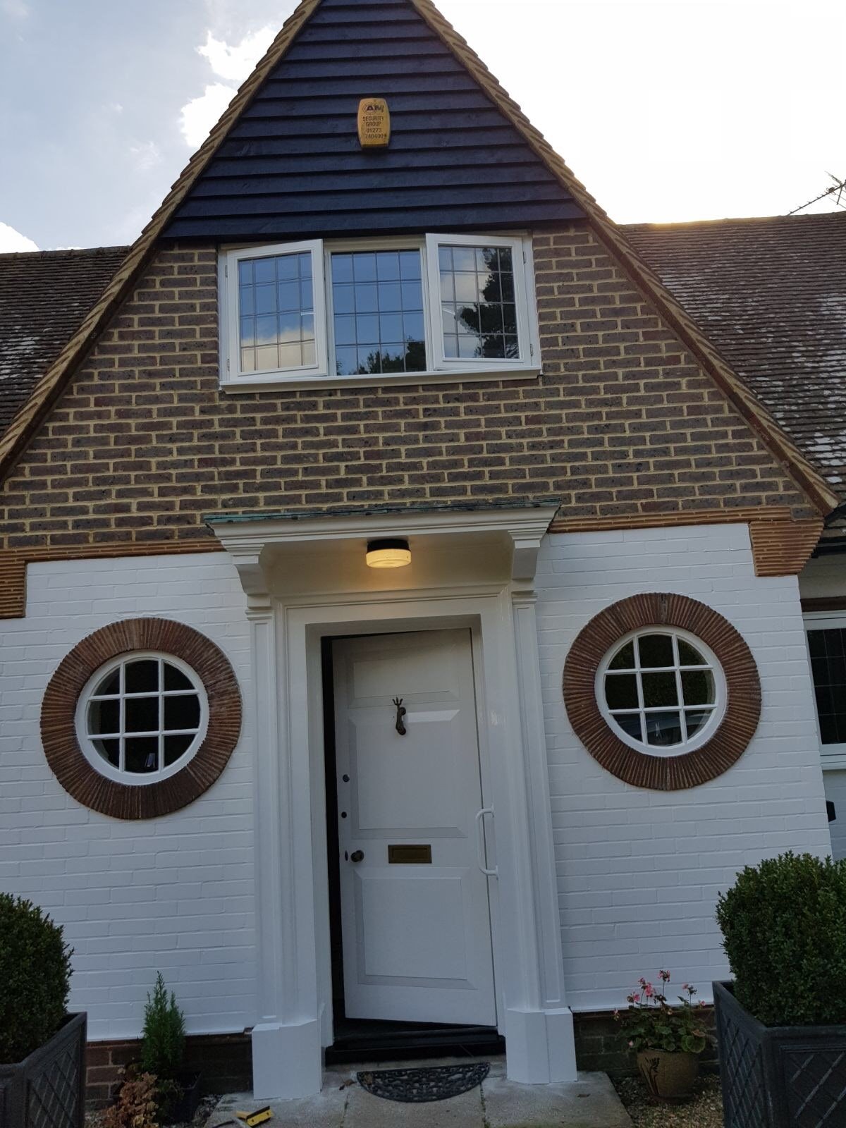 Front view of a house with a white door, two round windows with brown frames, and a brick facade on the upper part. The house has a peaked roof with dark shingles and a dormer window, with a security alarm mounted on the upper wall. Two small bushes 