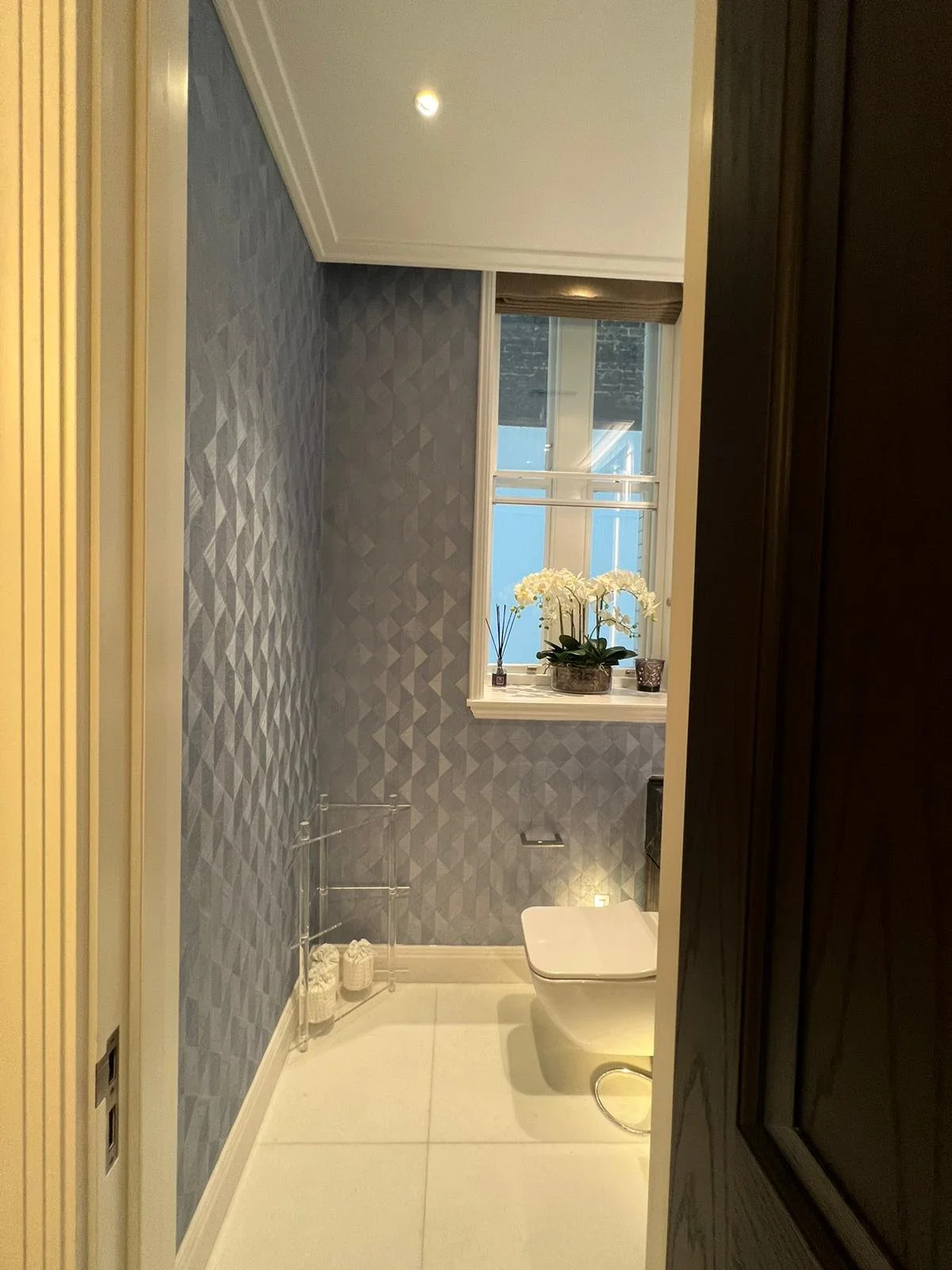 A modern bathroom with light-colored floor tiles, blue textured wallpaper, a window with a brown blind, a white toilet, and a white shelf with white orchids and decorative items.