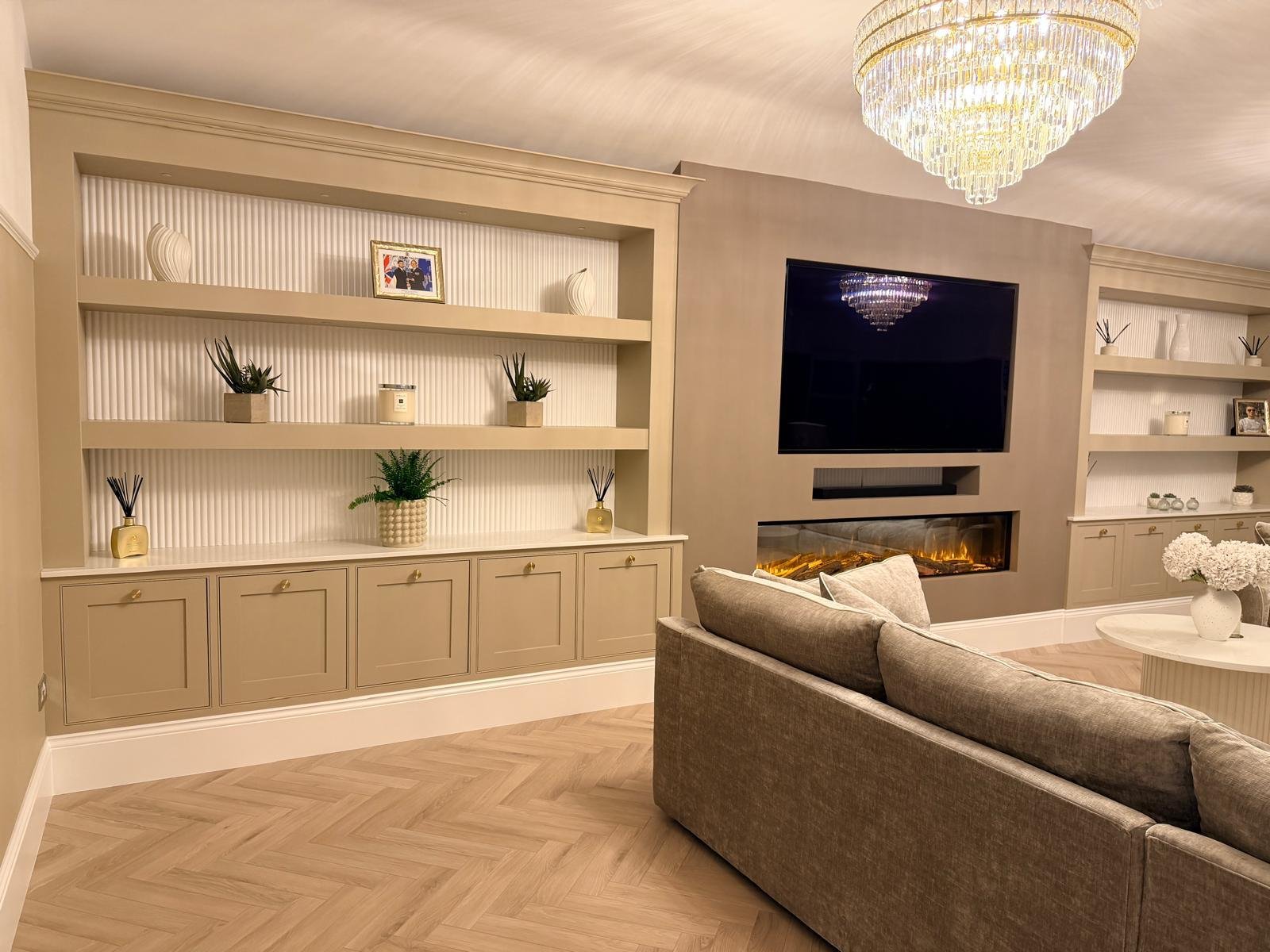 Living room with built-in shelves, wall-mounted flat-screen TV, an electric fireplace, beige sofa, chandelier, and decorative plants and items.