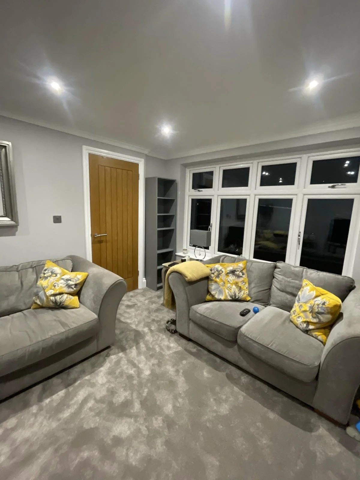 Living room with two gray sofas with yellow floral pillows and throws, a window with multiple panes, a small gray shelving unit, and a wooden door. There are remote controls, a blue ball, and other small objects on one sofa. The room has a gray carpe