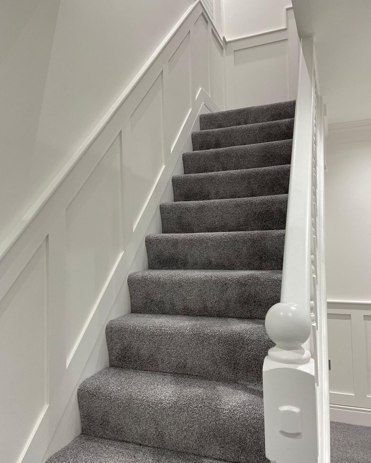 Indoor staircase with dark gray carpeted steps, white walls with wooden paneling, and a white handrail.
