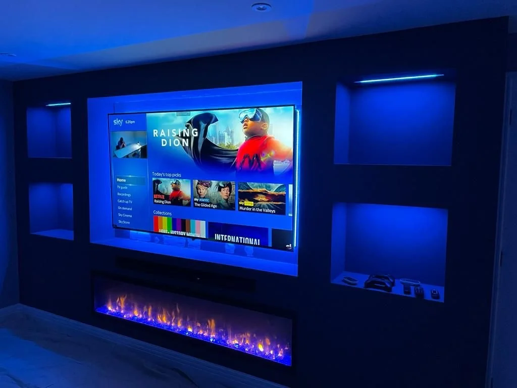 A living room features a large flat-screen TV displaying the Sky app with the show "Raising Dion" visible. The TV is mounted on a dark wall with blue accent lighting. Below the TV, there is a modern electric fireplace with blue flames. To the right o