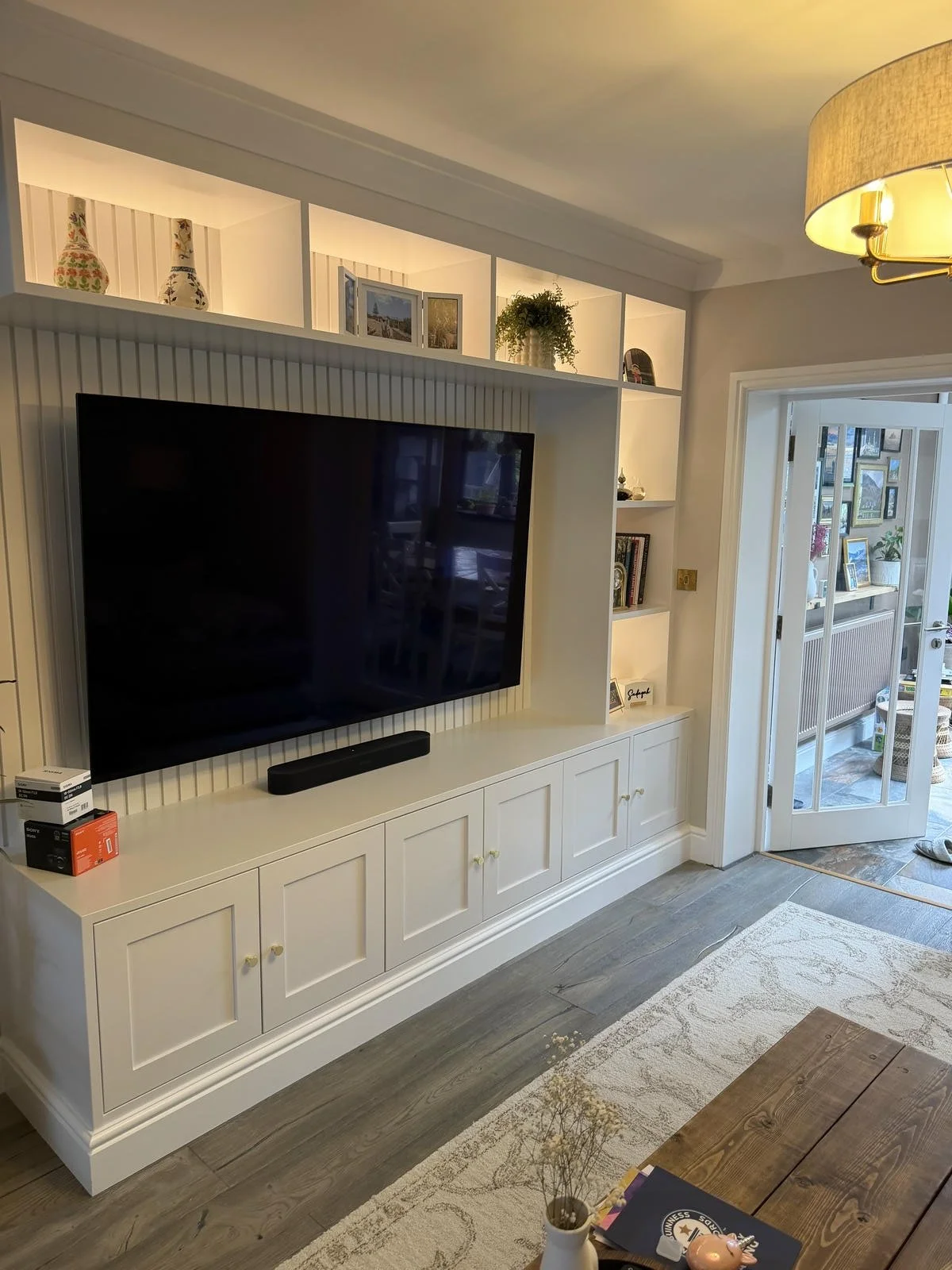 Living room with built-in white entertainment center, flat-screen TV, black soundbar, and decorative items on shelves. A doorway leading to a sunroom with furniture and artwork. Wooden table with a small potted plant and books.