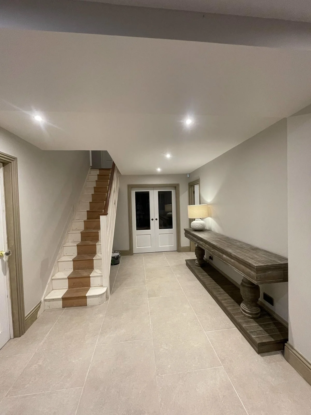 Interior of a modern home with beige tiled flooring, a staircase with wooden steps and white risers on the left, and a long wooden console table with turned legs on the right. There is a table lamp on the console table and two glass-paneled double doors at the back, with soft lighting in the room.