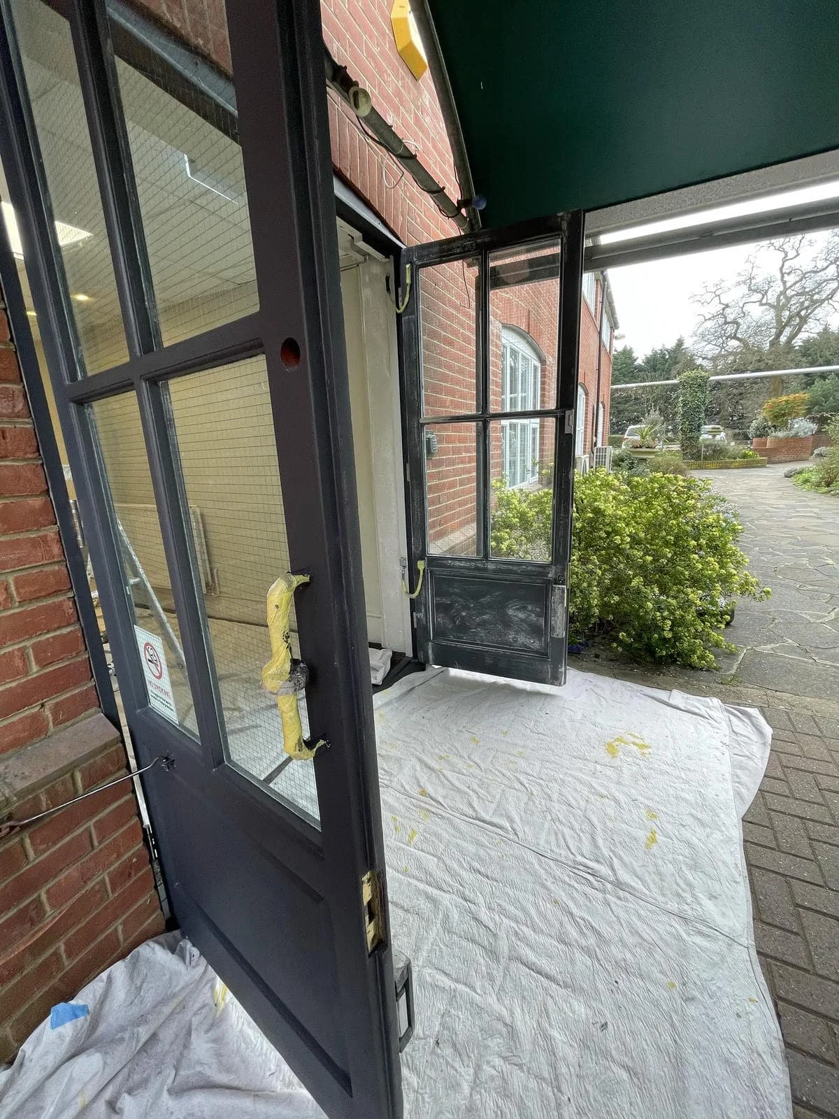 A black exterior door with multiple glass panels is open, revealing an interior with beige walls and a window. The door has yellow protective padding on the handle and is placed on a white tarp outside a brick building, with a garden and paved path i