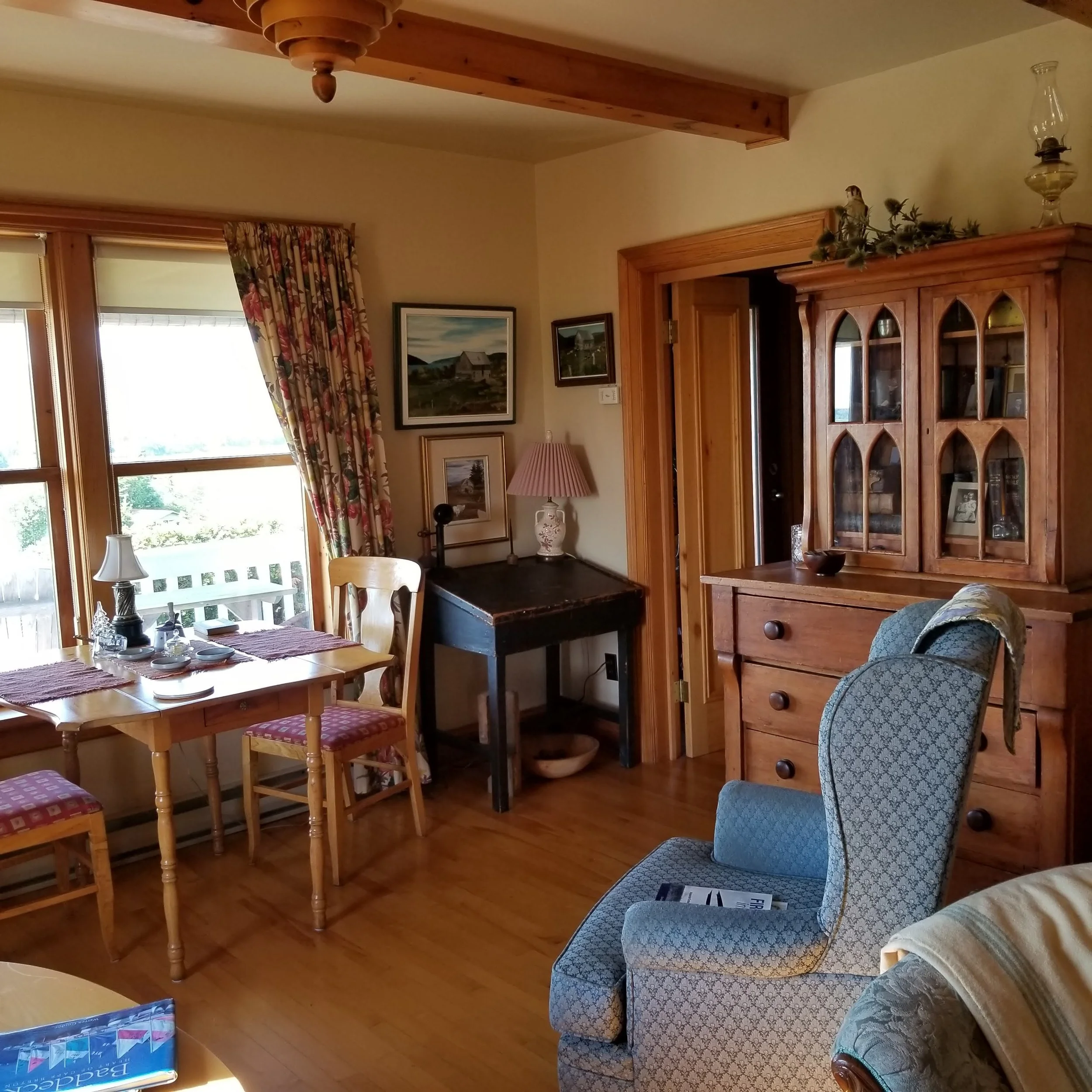 A cozy living room with wooden accents, two large windows with floral curtains, a wooden dining table with chairs, antique cabinet, several framed paintings on the wall and a blue patterned armchair.