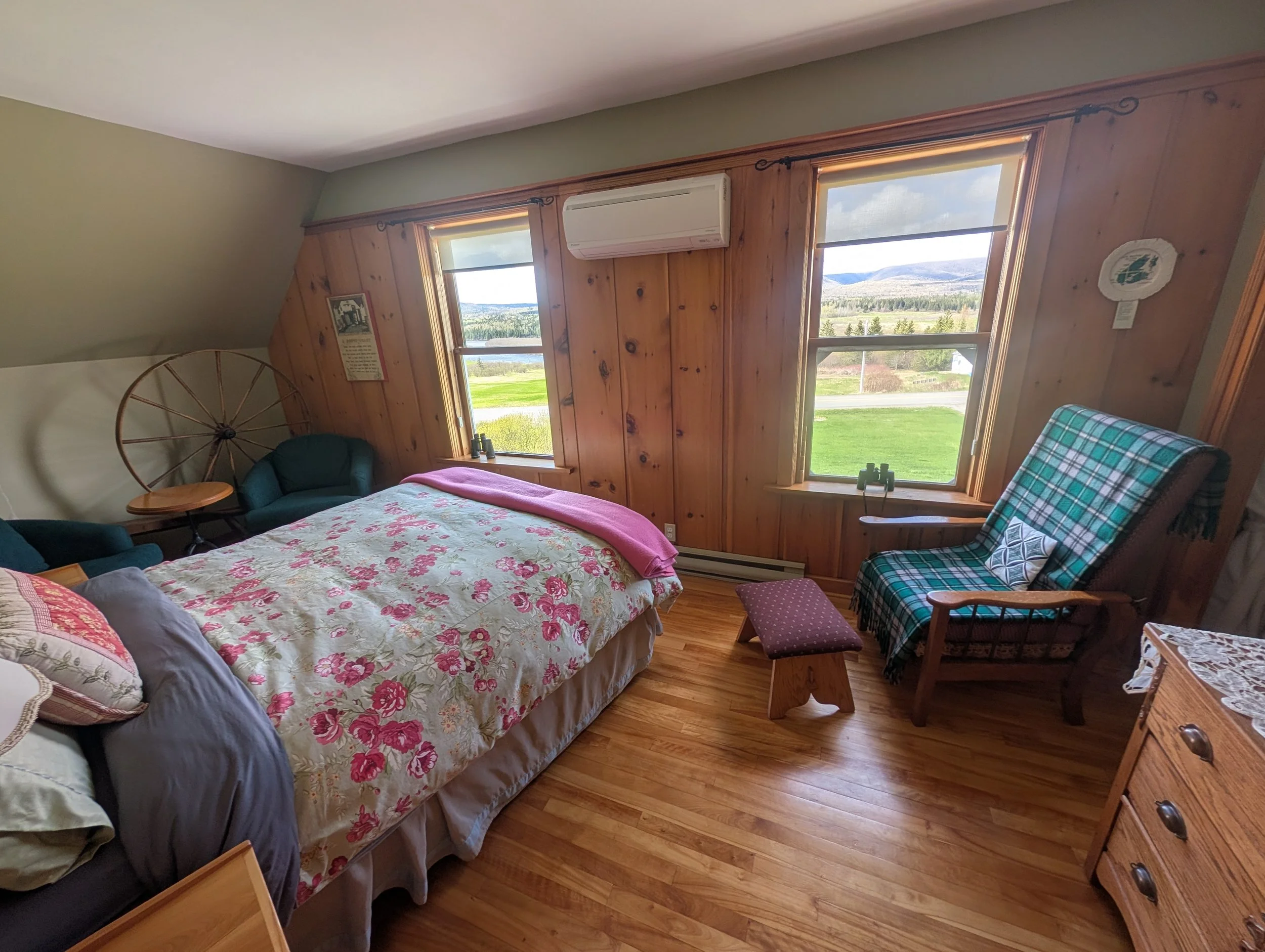 A cozy bedroom with wood-paneled walls and large windows showing a scenic landscape with trees and mountains. The bed has a floral coverlet and pillows, a plaid armchair, and a small wooden side table. There are binoculars on the window sill, an air conditioning unit, and framed pictures on the walls.