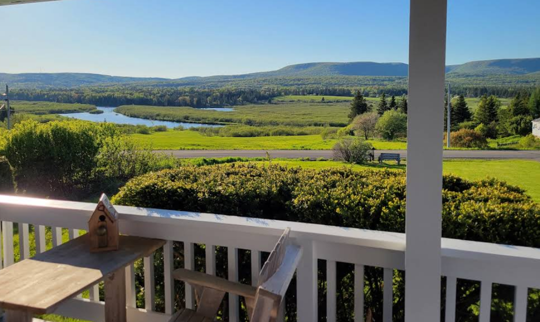 View of a lush green landscape with a winding river, rolling hills, and trees, seen from a porch with a white railing and a birdhouse on a wooden table in the foreground.