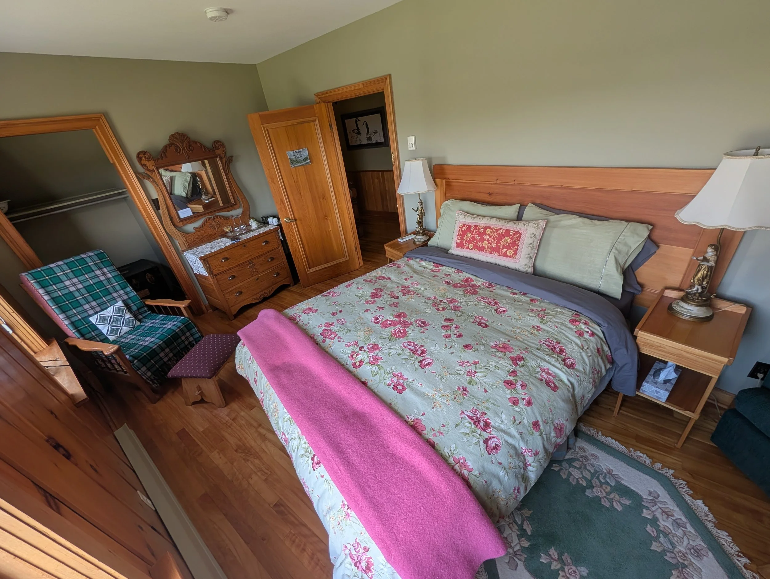 A cozy bedroom with a wooden bed, floral bedspread, and pillows, next to a nightstand with a vintage lamp. There is an armchair with island tartan cloth, a wooden dresser with a mirror, and a closet. The room has wooden flooring and lightgreen walls.