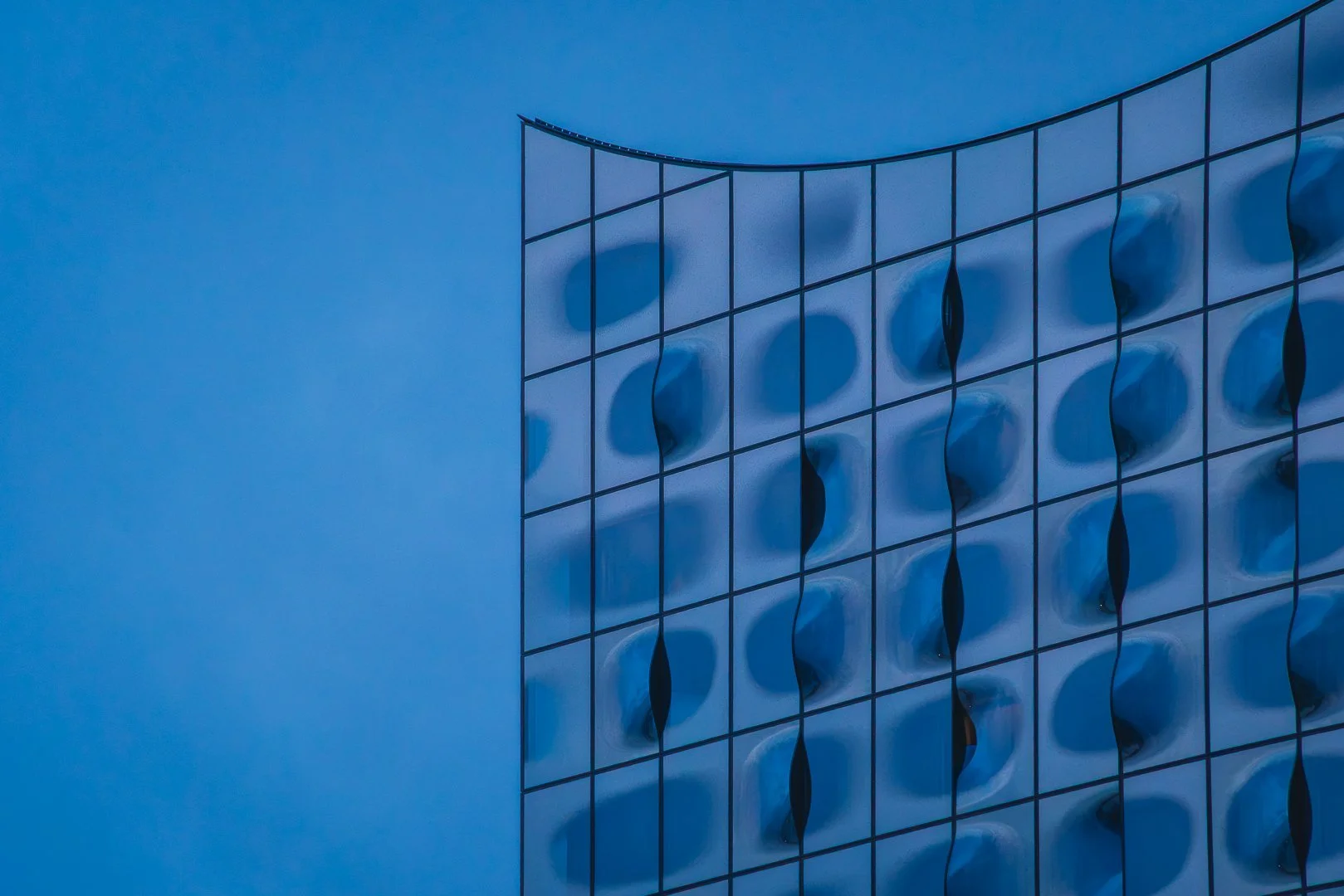 Abstract blue-hour photograph of Hamburg’s Elbphilharmonie with a curved glass facade entering a large field of blue negative space.