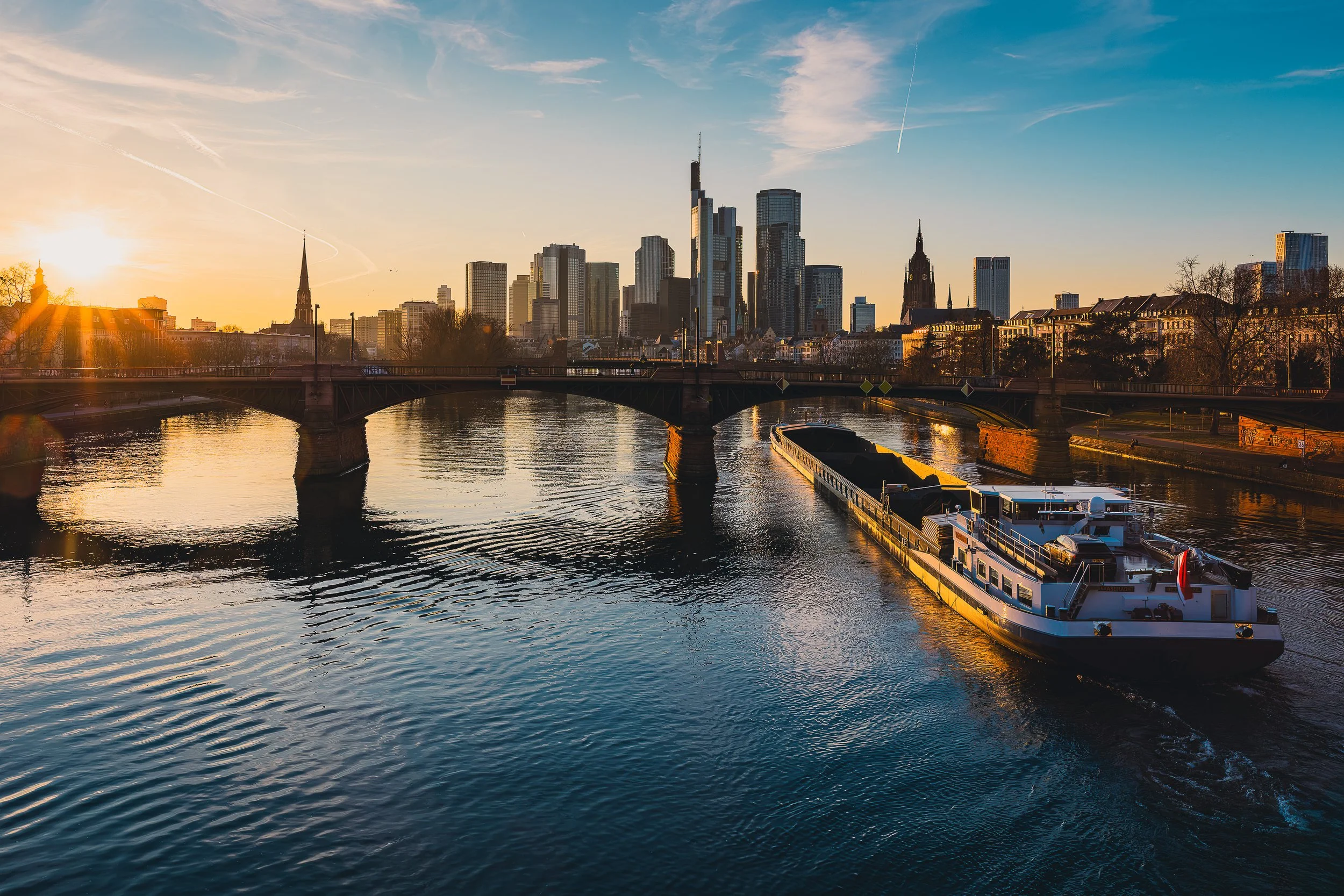 Frankfurt skyline at sunset with Flößerbrücke and a cargo ship on the Main River