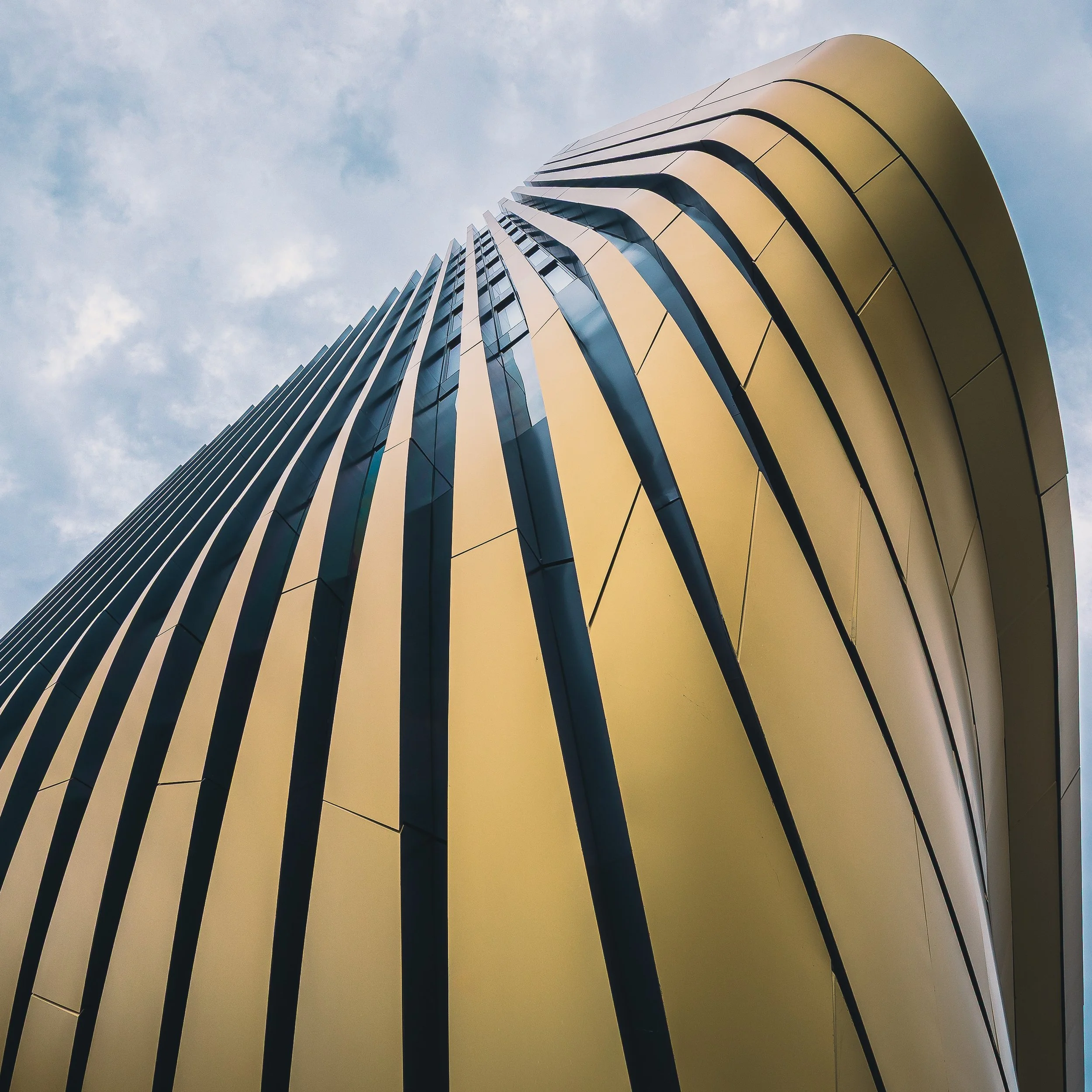 Curved golden facade with dark vertical lines rising into a cloudy sky in Prague